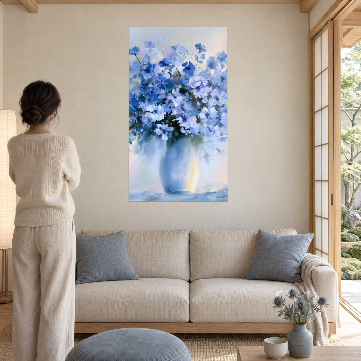 Tableau bouquet de bleuets bleu cobalt et blanc crème dans un vase bleuté, femme de dos, au-dessus d'un canapé dans un salon lumineux