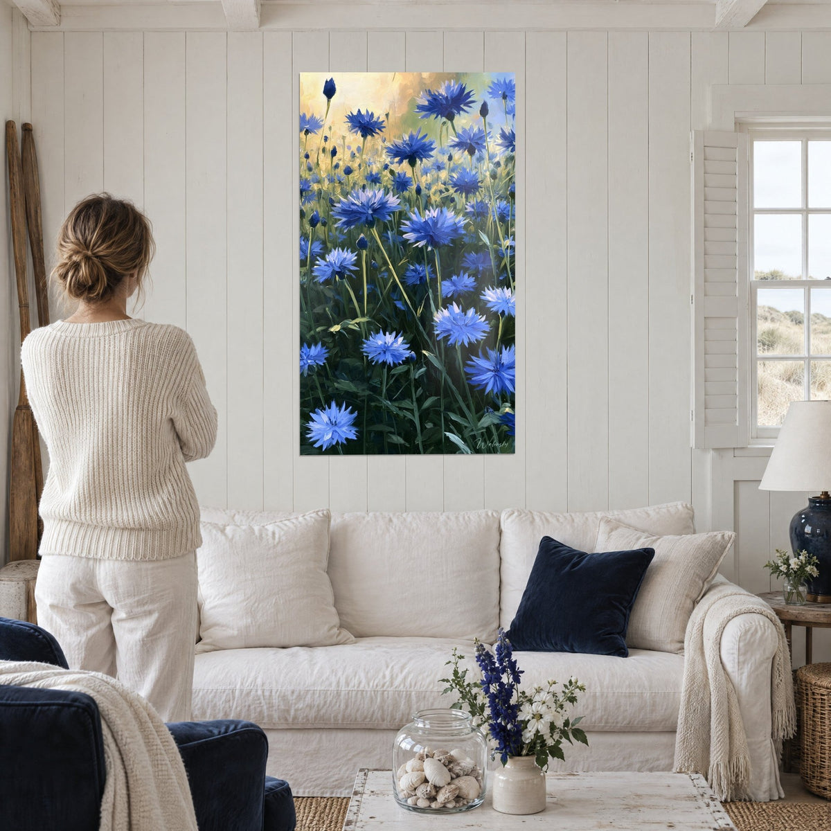 Tableau champ de bleuets bleu cobalt sous un ciel jaune doré, vu d'en bas, femme de dos, au-dessus d'un canapé dans un salon lumineux