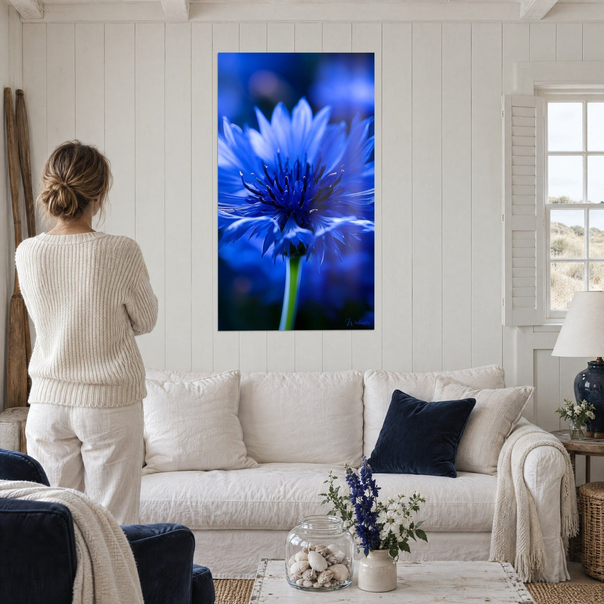 Tableau bleuet électrique aux pétales frangés et tige verte, une femme de dos au-dessus d'un canapé dans un salon lumineux