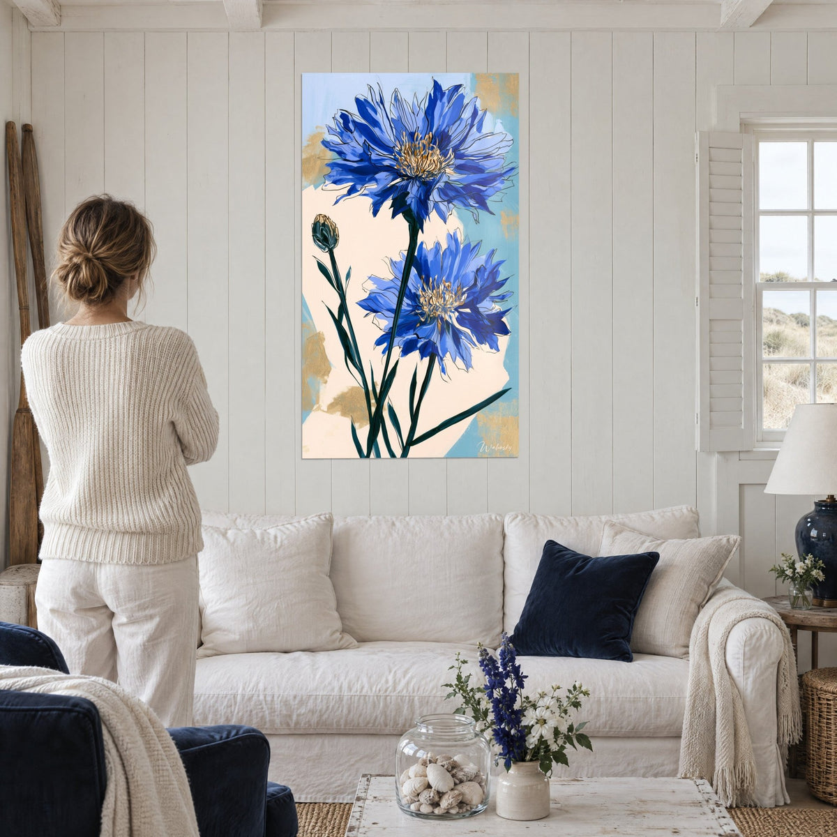 Schilderij met indigokleurige korenbloemen en saffraangele hartjes op een crèmekleurige achtergrond, boven een bank in een lichte woonkamer