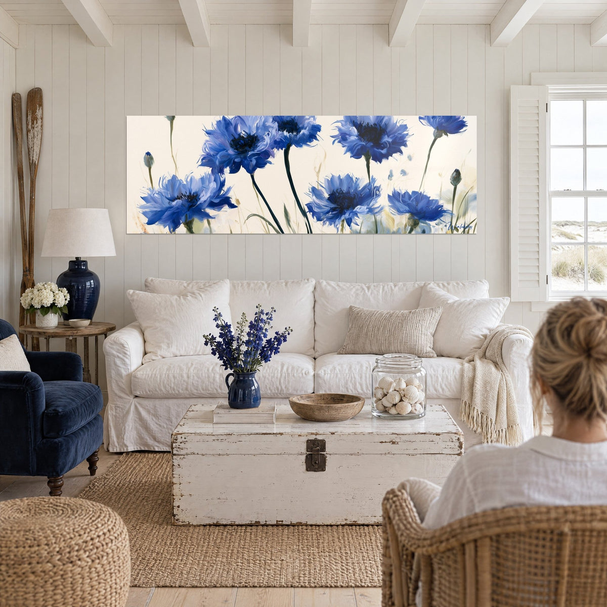 Tableau bleuets bleu cobalt aux pétales frangés sur fond ivoire, deux personnes de dos, au-dessus d'un canapé dans un salon lumineux