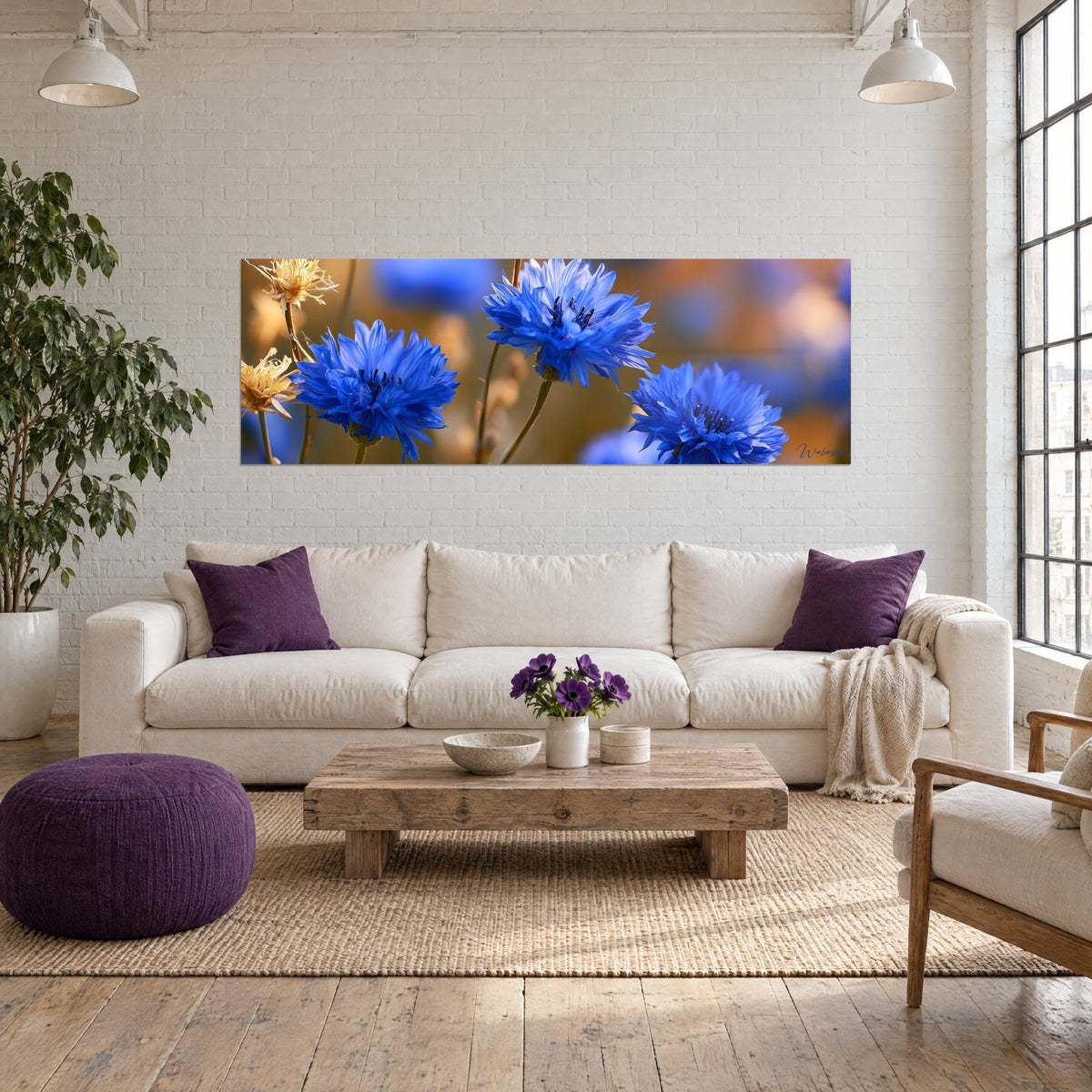 Gemälde Kornblumen in leuchtendem Blau mit gefransten Blütenblättern vor unscharfem Rehbraun, über einem Sofa in einem hellen Wohnzimmer