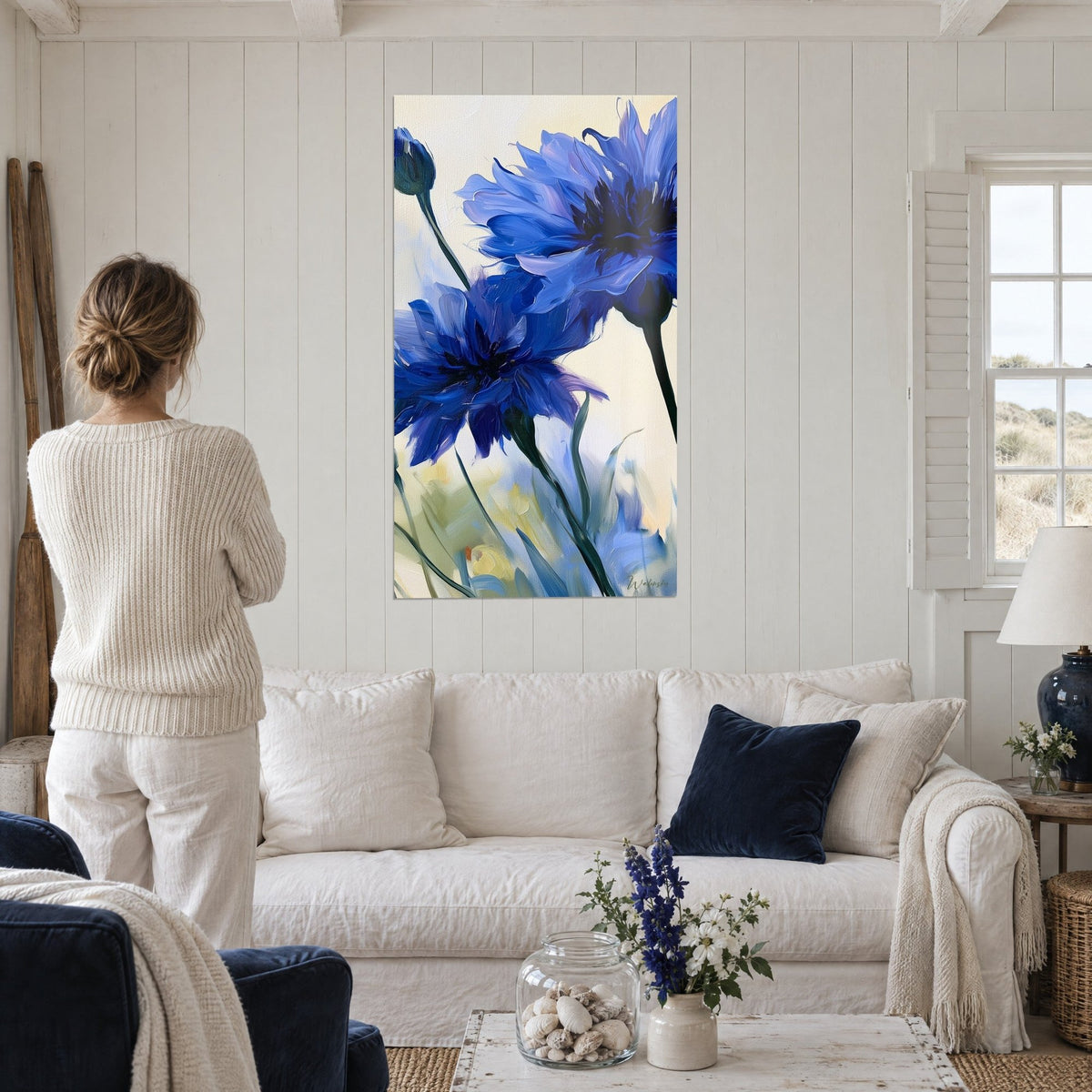 Quadro di fiordalisi indaco dai petali stropicciati, sfondo avorio, steli verde scuro, visto di spalle sopra un divano in un soggiorno luminoso