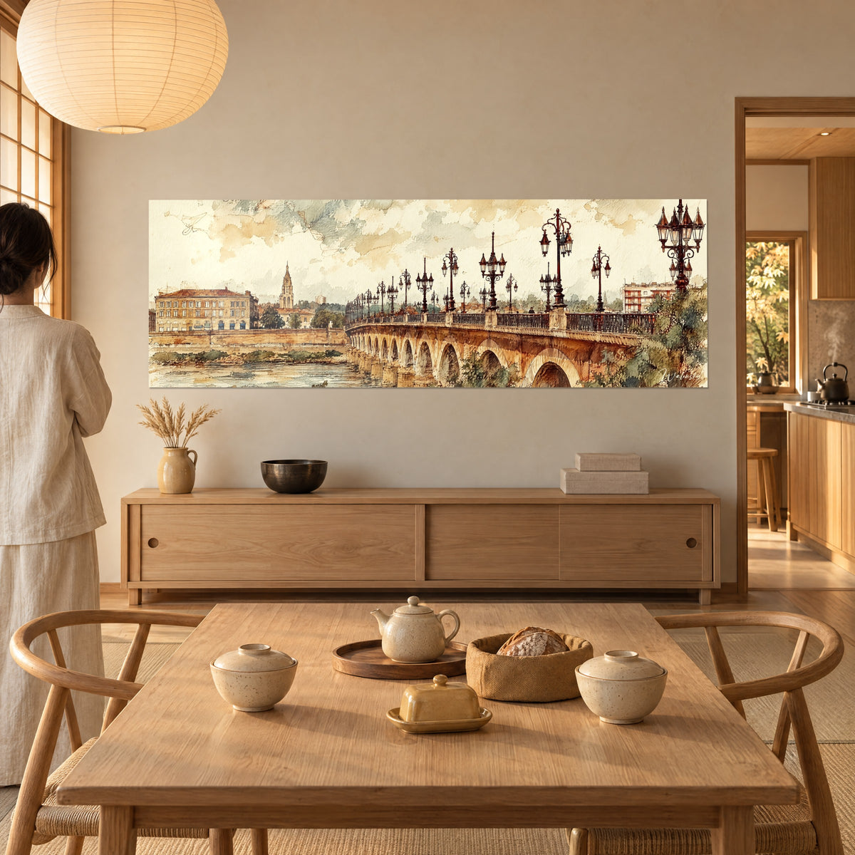 Cuadro río azul grisáceo bajo un puente de arcos ocre, linternas oscuras alineadas, admirado por una mujer de espaldas junto a una mesa de comedor