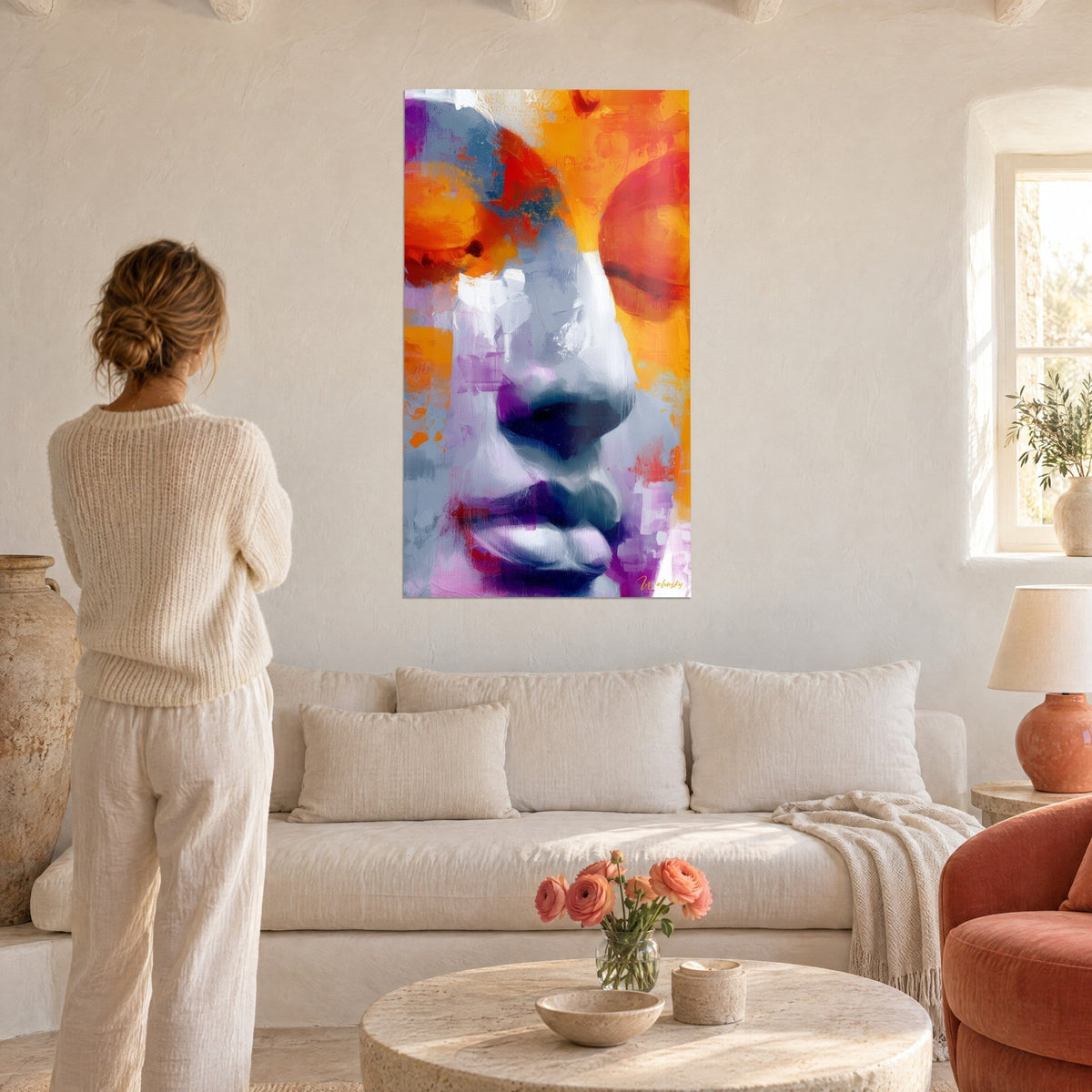 Tableau visage de bouddha en gros plan, paupières orange flamboyant et lèvres violettes, une femme de dos, au-dessus d'un canapé dans un salon lumineux