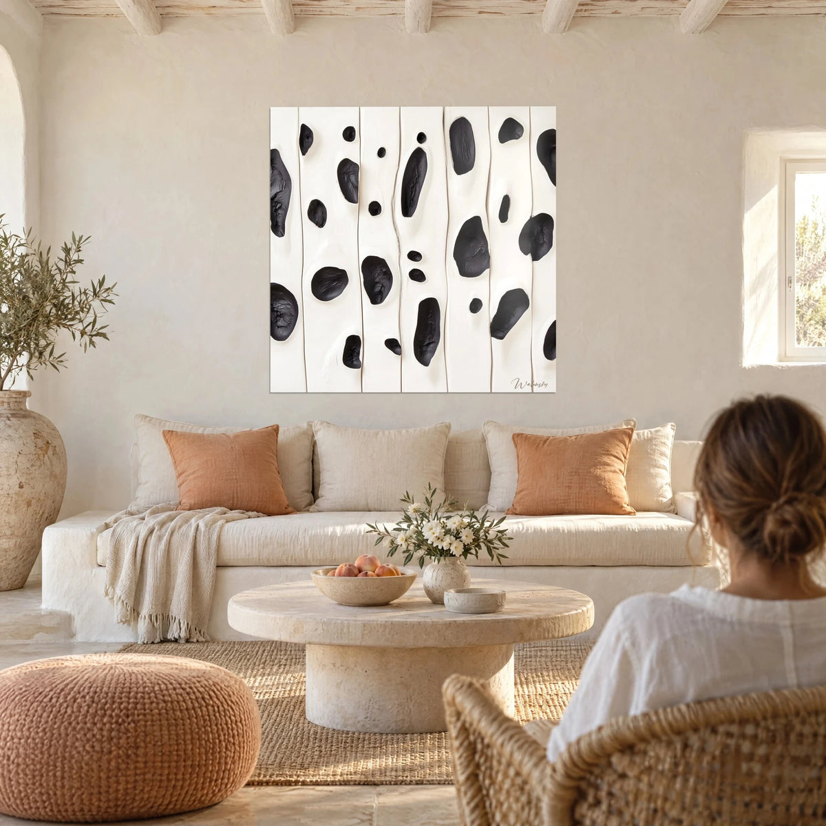 Schilderij van gestileerde berken met zwarte inktvlekken op een crèmekleurige achtergrond, een vrouw met de rug naar de kijker boven een bank in een lichte woonkamer