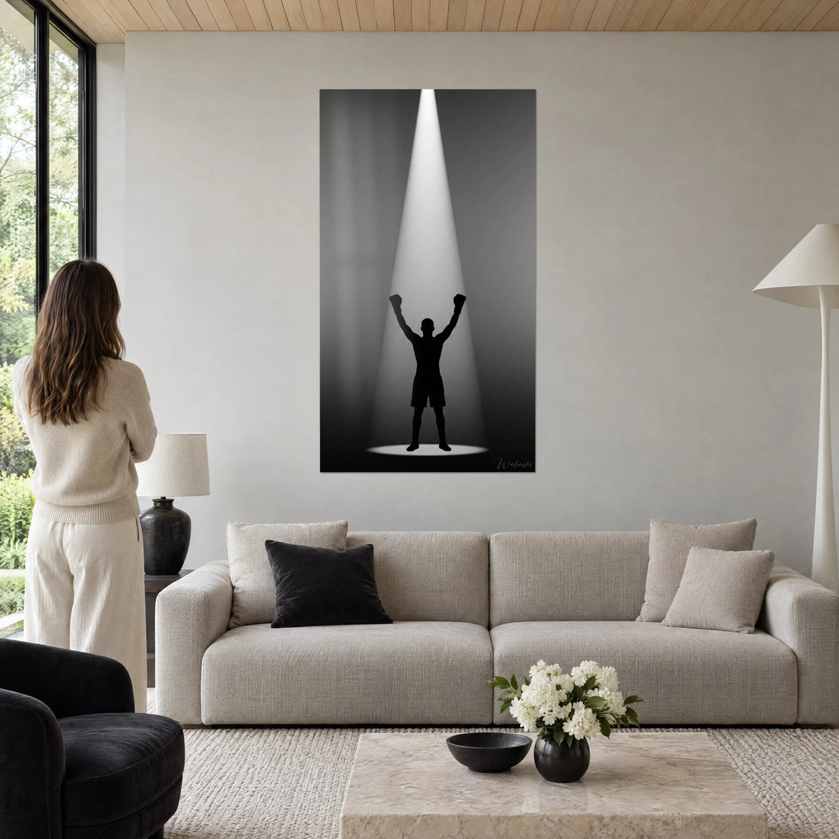 Quadro pugile in sagoma nera, guantoni alzati sotto un fascio bianco su sfondo grigio, donna di spalle, sopra un divano in un soggiorno luminoso