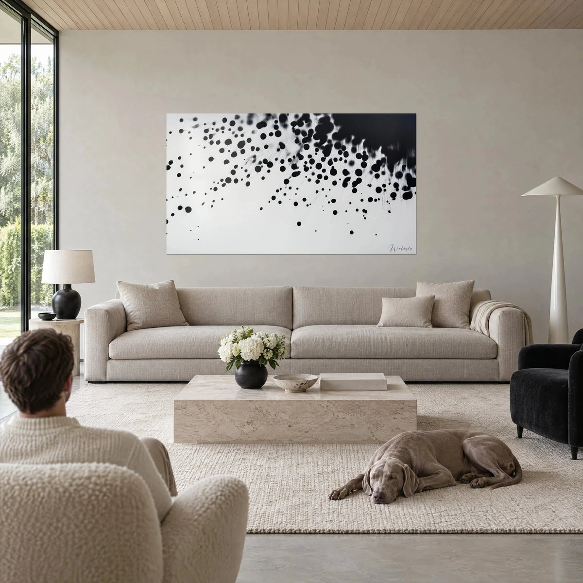 Wall art with rain of black ink drops on white background, two people seen from behind and a Weimaraner, above a sofa in a bright living room