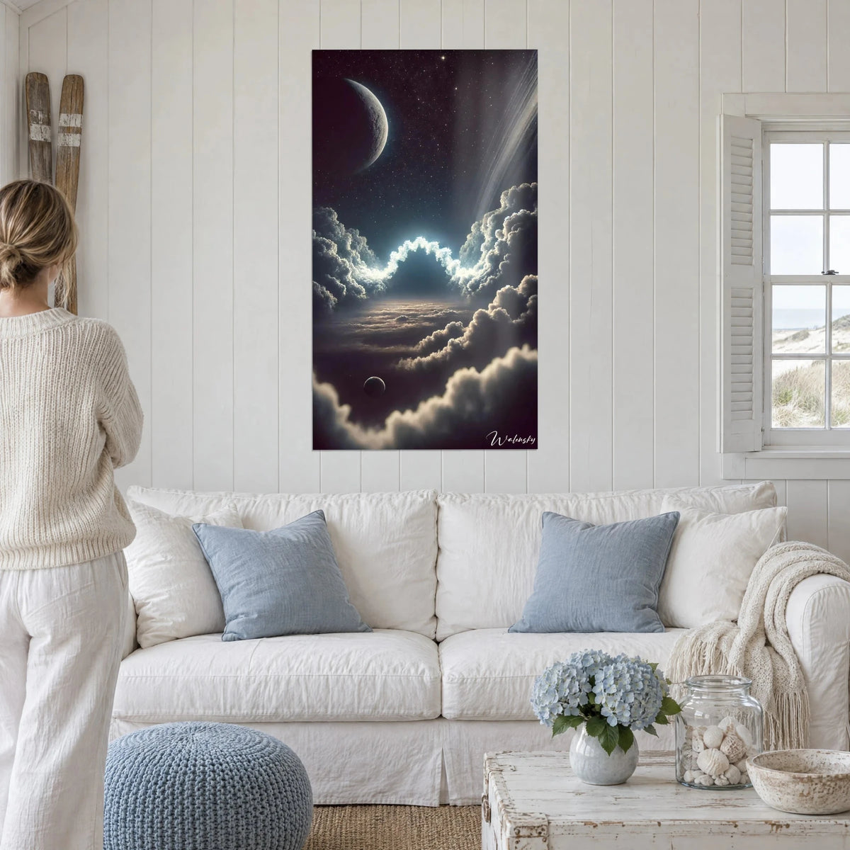 Tableau ciel étoilé aux nuages éclairés d'un halo bleu glacé et lune pâle, femme de dos, au-dessus d'un canapé dans un salon lumineux