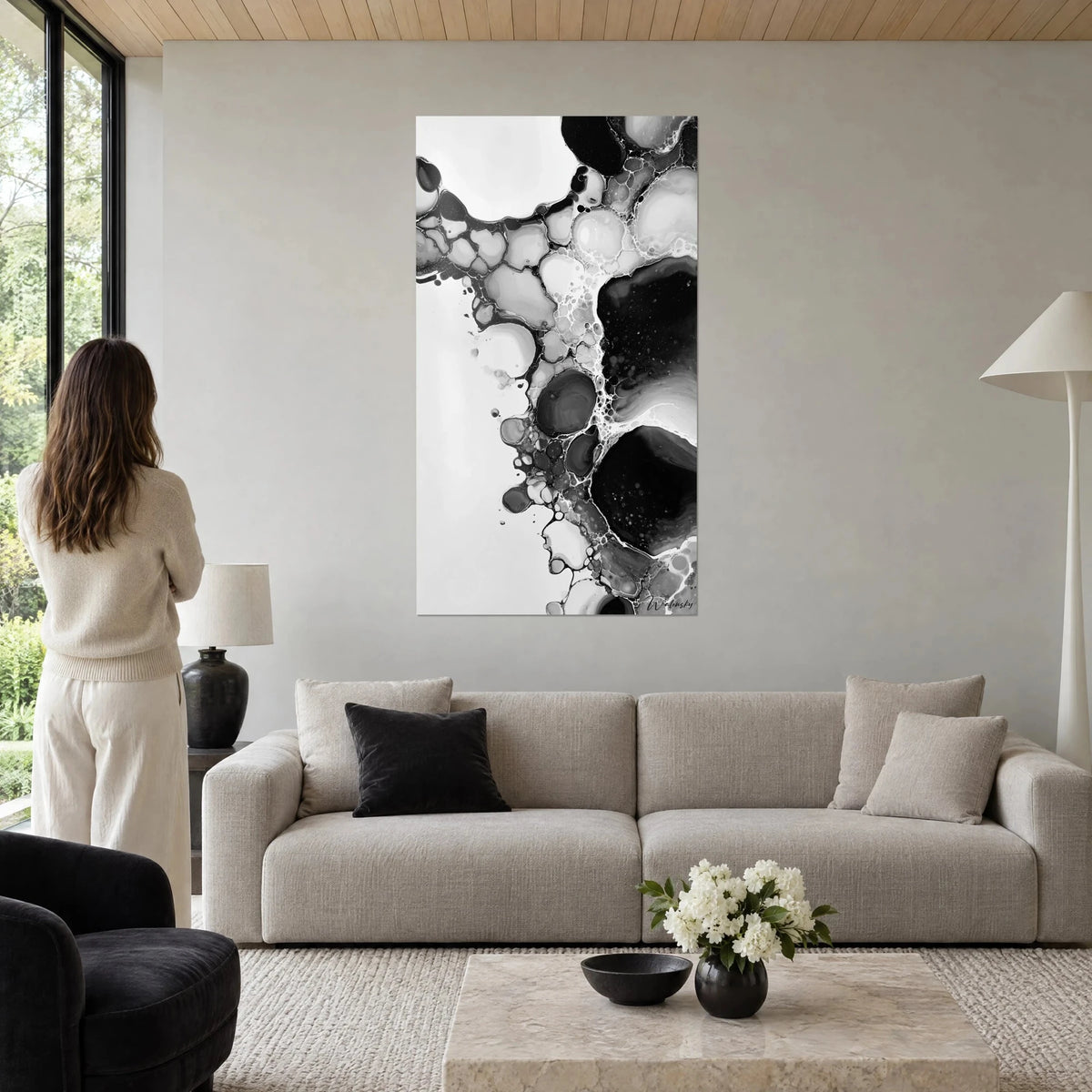 Wall art with black and grey ink cells on a white background, a woman seen from behind gazes at it above a sofa in a bright living room