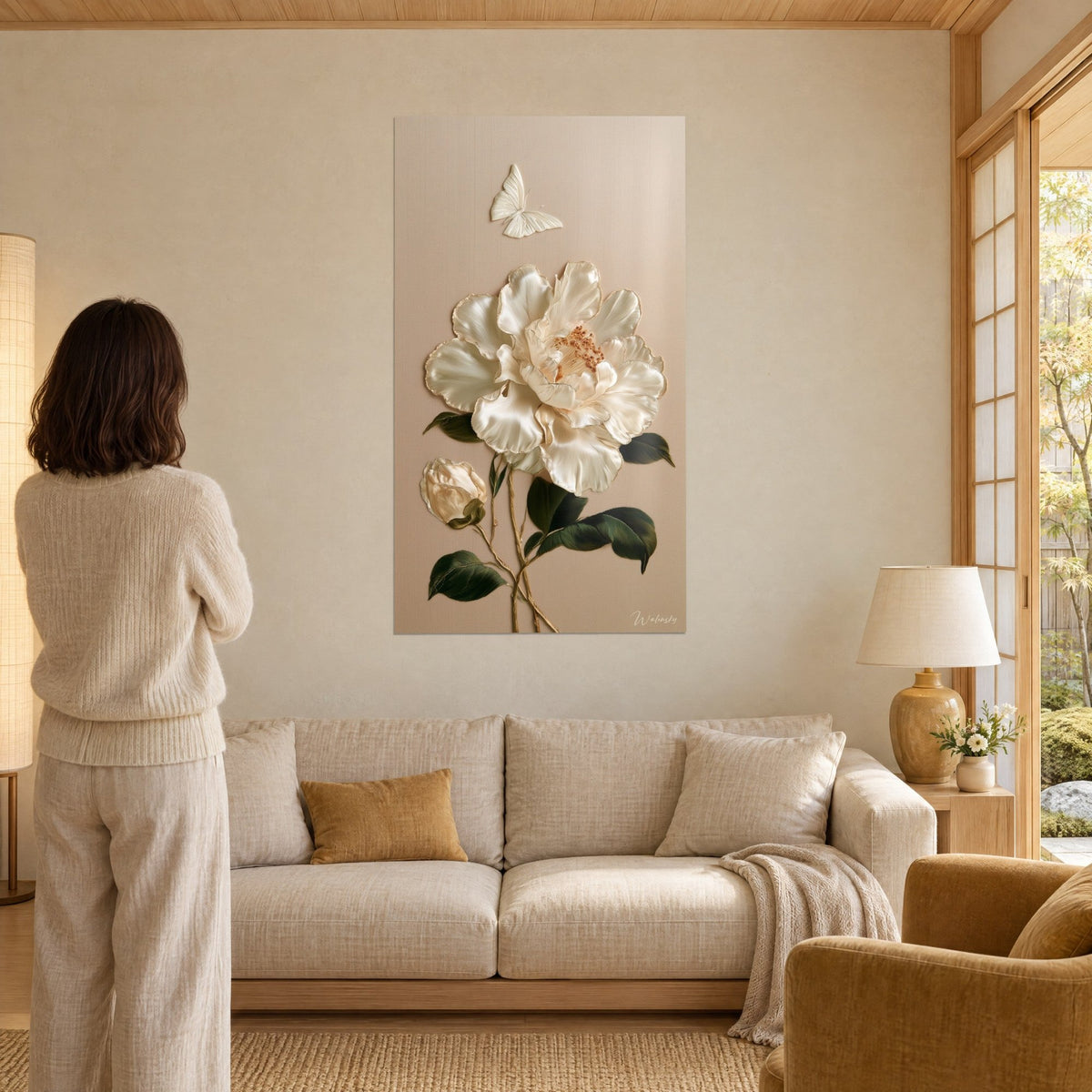 Cuadro camelia de satén marfil y mariposa nacarada sobre fondo beige, mujer de espaldas, encima de un sofá en un salón luminoso
