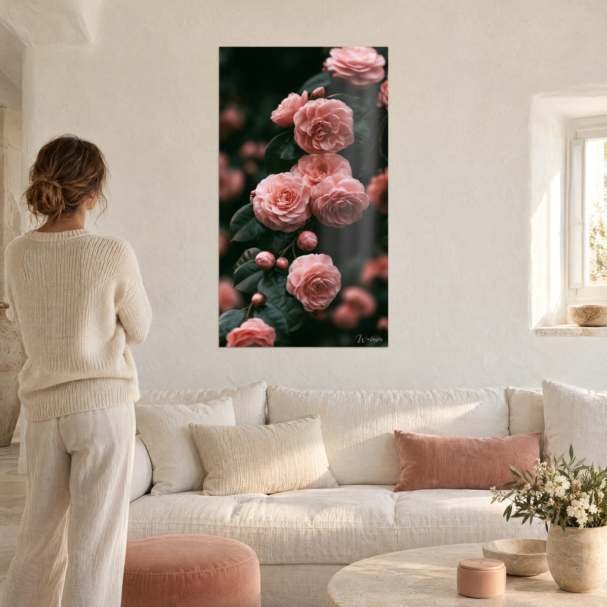 Quadro con camelie color salmone in piena fioritura su fogliame verde scuro, donna di spalle con maglione color crema, sopra un divano in un soggiorno luminoso