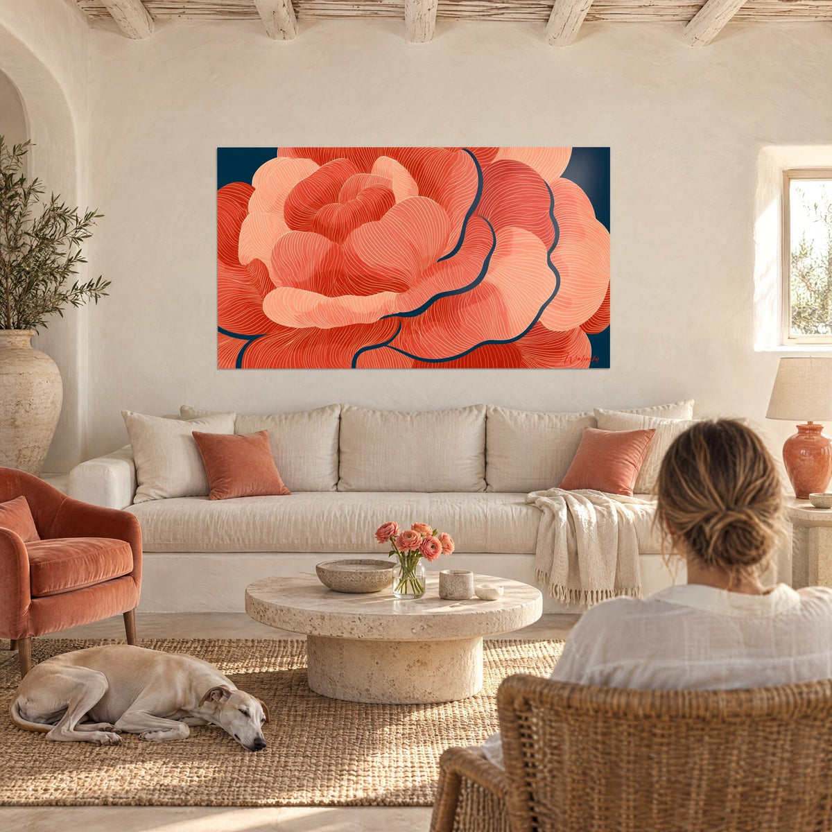 Quadro camelia corallo dai petali ondulati su fondo blu navy, donna di spalle e levriero, sopra un divano in un soggiorno luminoso
