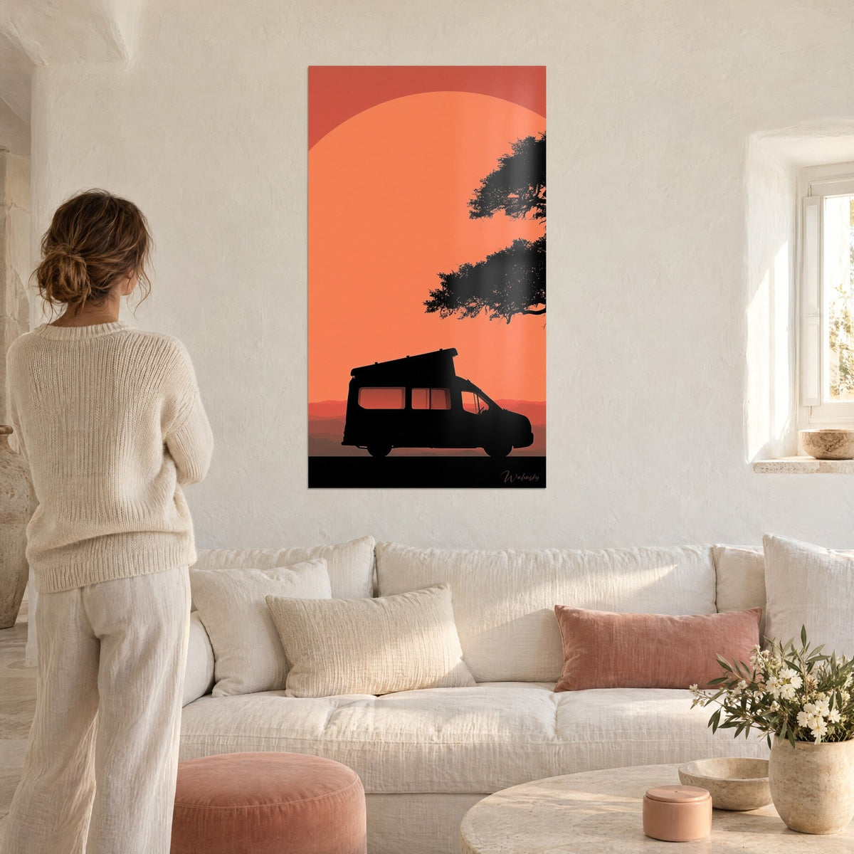 Wall art camper van in black silhouette under a parasol pine, huge orange sun, woman seen from behind, above a sofa in a bright living room