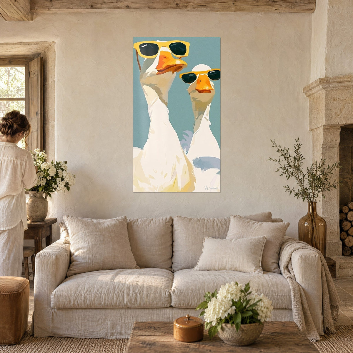 Schilderij twee witte ganzen met gele zonnebril op een blauwgroene achtergrond, een vrouw met haar rug naar de kijker, boven een bank in een lichte woonkamer
