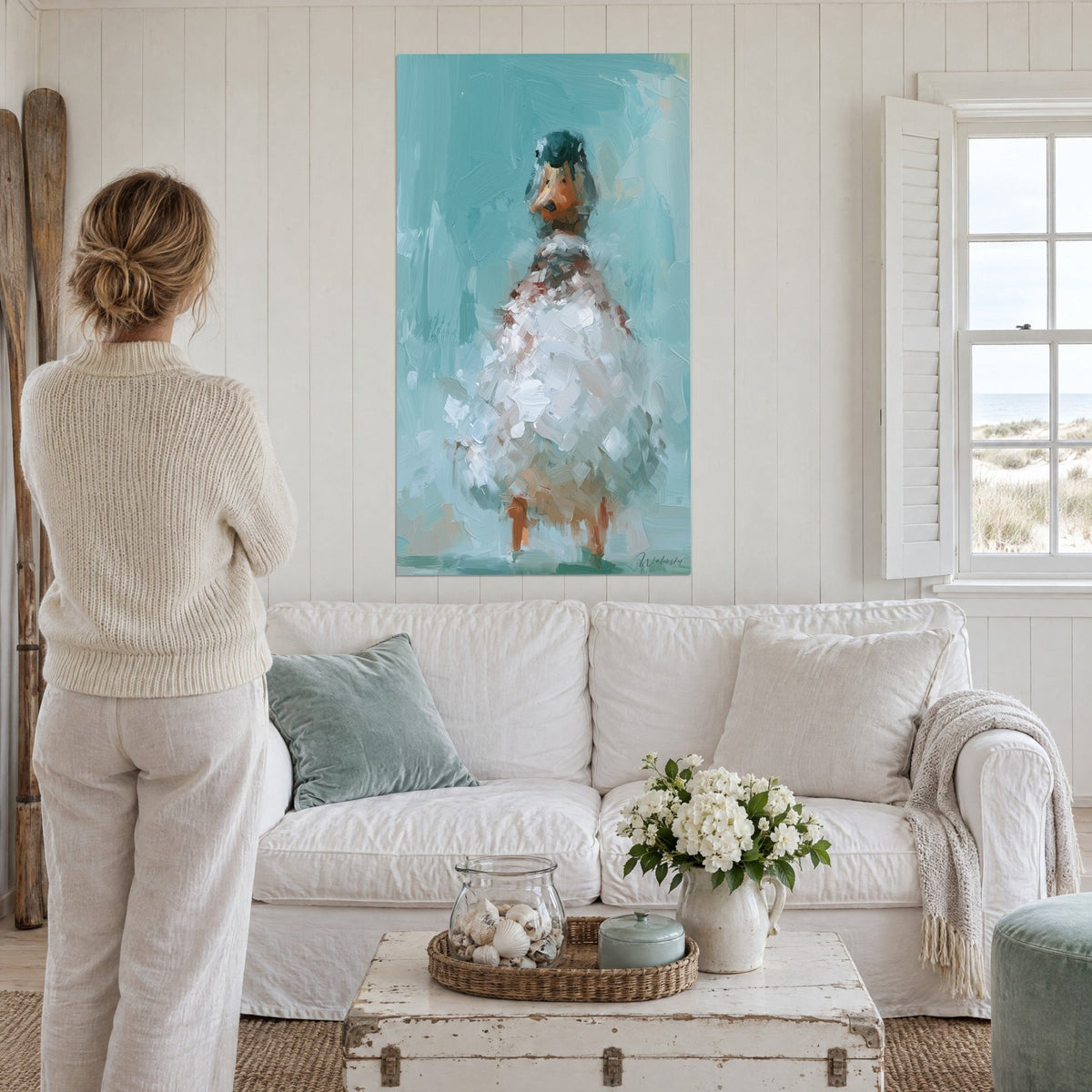 Quadro di un'anatra bianca dal piumaggio di tocchi madreperlacei, sfondo blu petrolio, una donna di spalle, sopra un divano in un soggiorno luminoso