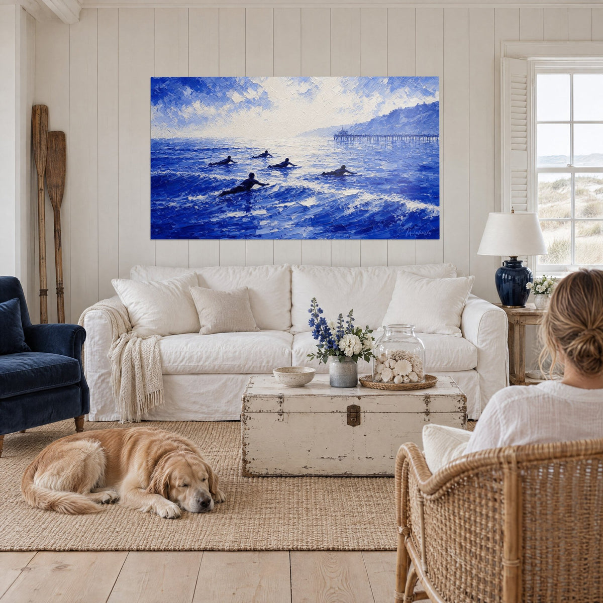 Wall art surfers lying on their boards paddling toward the cobalt and white swell, distant jetty, above a sofa in a bright living room