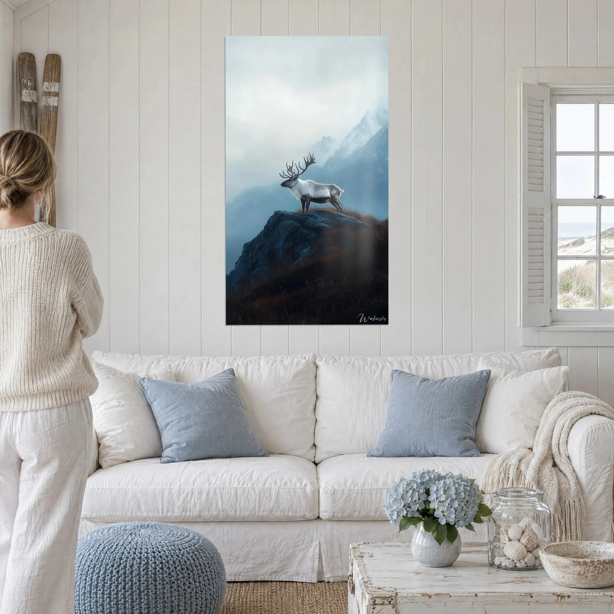 Gemälde mit einem weißen Karibu auf einem nachtblauen Felsen, grauer Nebel und verschneite Gipfel, über einem Sofa in einem hellen Wohnzimmer