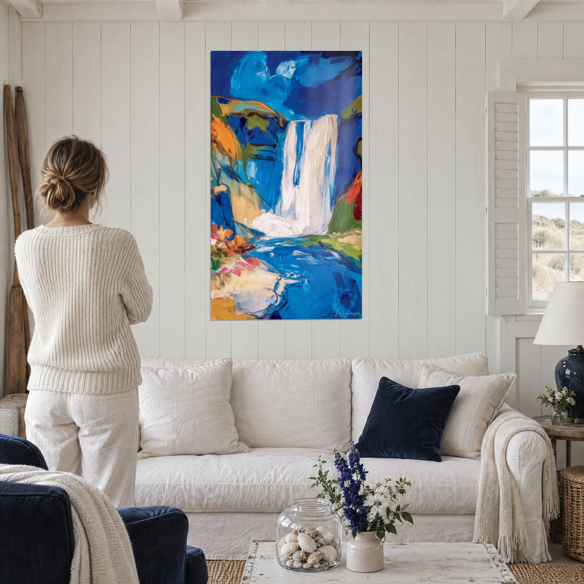 Wall art white waterfall in thick impasto on a cobalt blue and ochre background, woman seen from behind, above a sofa in a bright living room