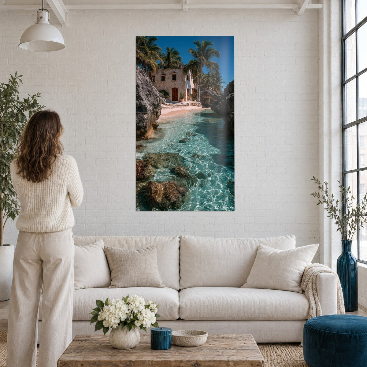 Wall art of a tropical cove with turquoise waters, pale stone house and palm trees, on a white brick wall above a sofa in a bright living room