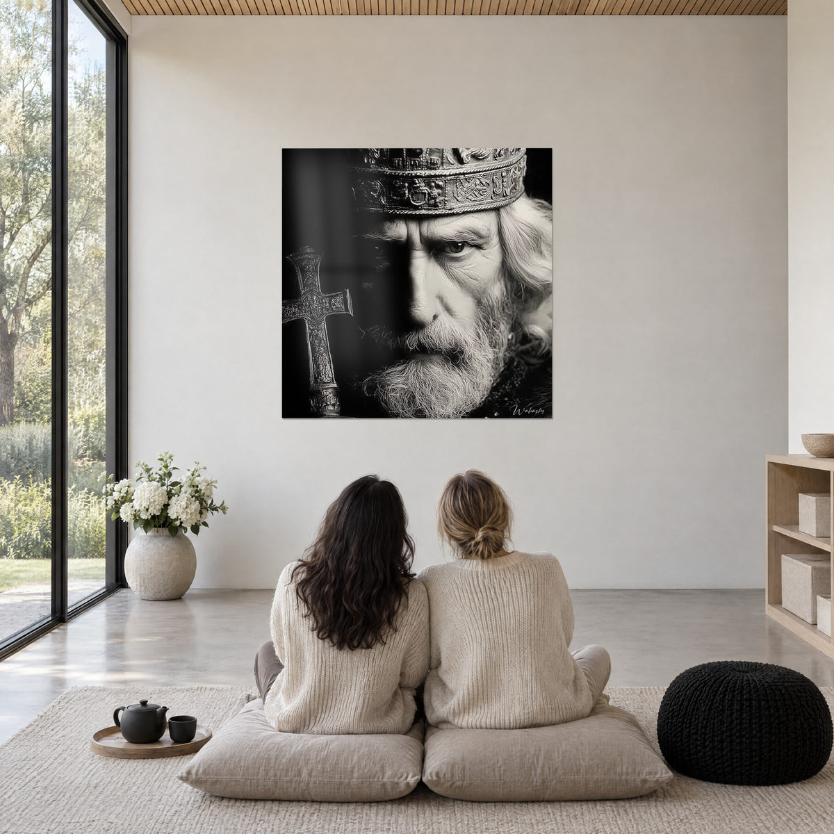 Cuadro de un anciano rey de barba poblada, corona cincelada, blanco y negro, pared blanca, dos mujeres de espaldas sentadas sobre cojines
