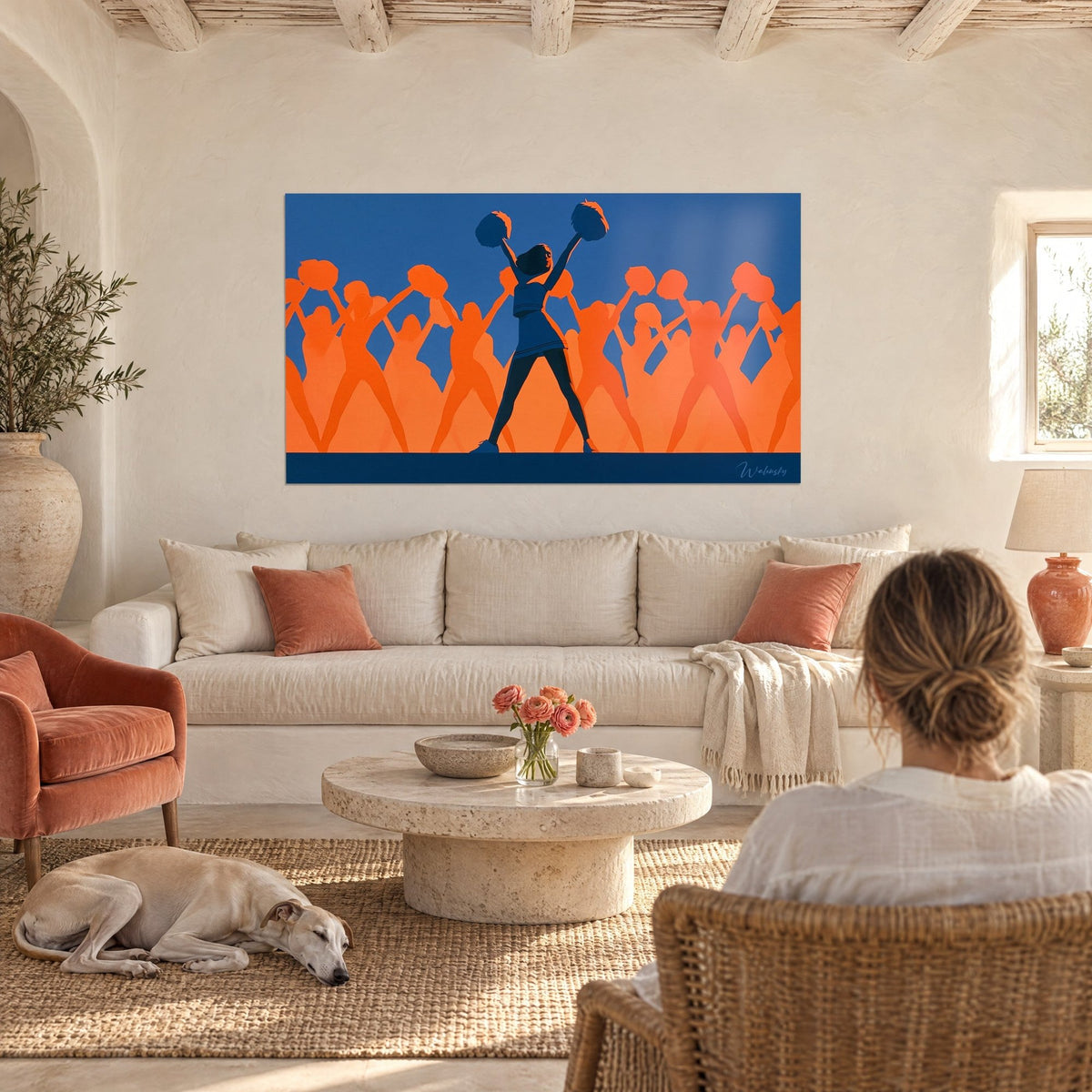 Quadro cheerleader con le braccia alzate, pon pon blu notte e sagome arancioni, una donna di spalle sopra un divano in un soggiorno luminoso