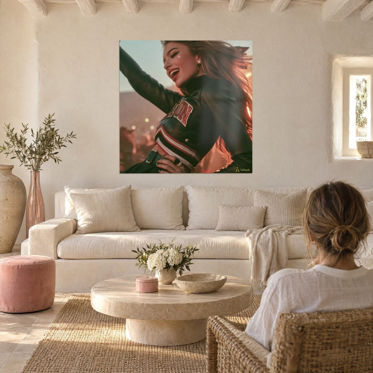 Wall art of a laughing cheerleader in a black bomber jacket, arm raised, pink and cream sky, above a sofa in a bright living room