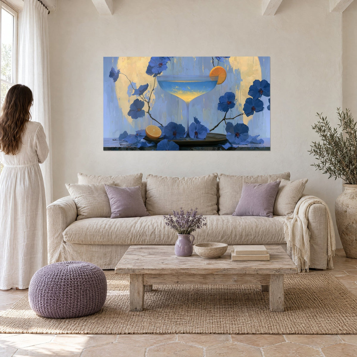 Schilderij cocktailcoupe met blauw en gouden verloop, indigo bloemen, sinaasappelschijfje, vrouw van achteren, boven een bank in een lichte woonkamer