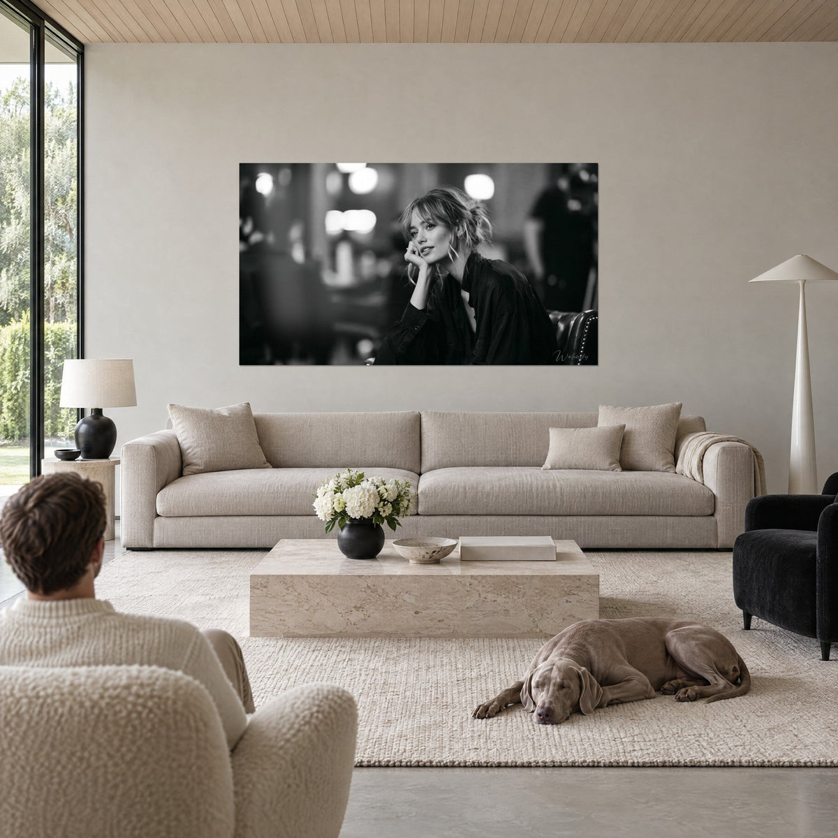 Wall art of a pensive young woman in black and white, bangs and loose bun, cheek resting on her hand, Weimaraner lying nearby, above a sofa in a bright living room