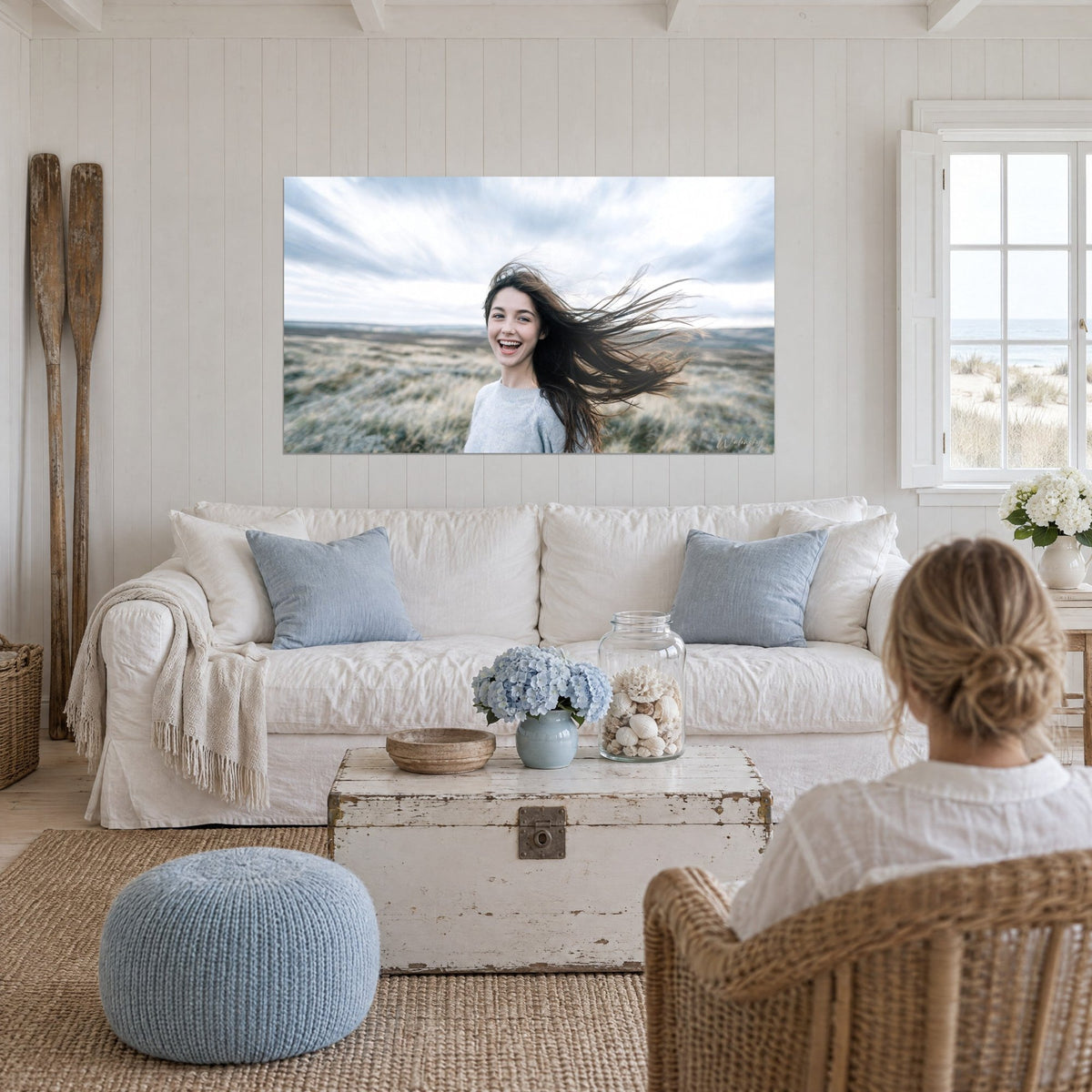 Wall art of a laughing young woman with brown hair whipped by the wind, pearl-grey sweater, cloudy sky, above a sofa in a bright living room