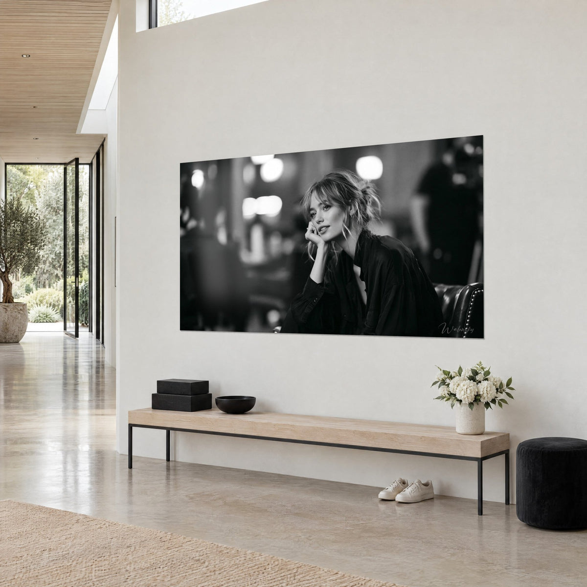 Wall art portrait with a dreamy gaze, wispy strands and oversized black shirt, dark leather armchair, cream wall and wooden bench, three-quarter view