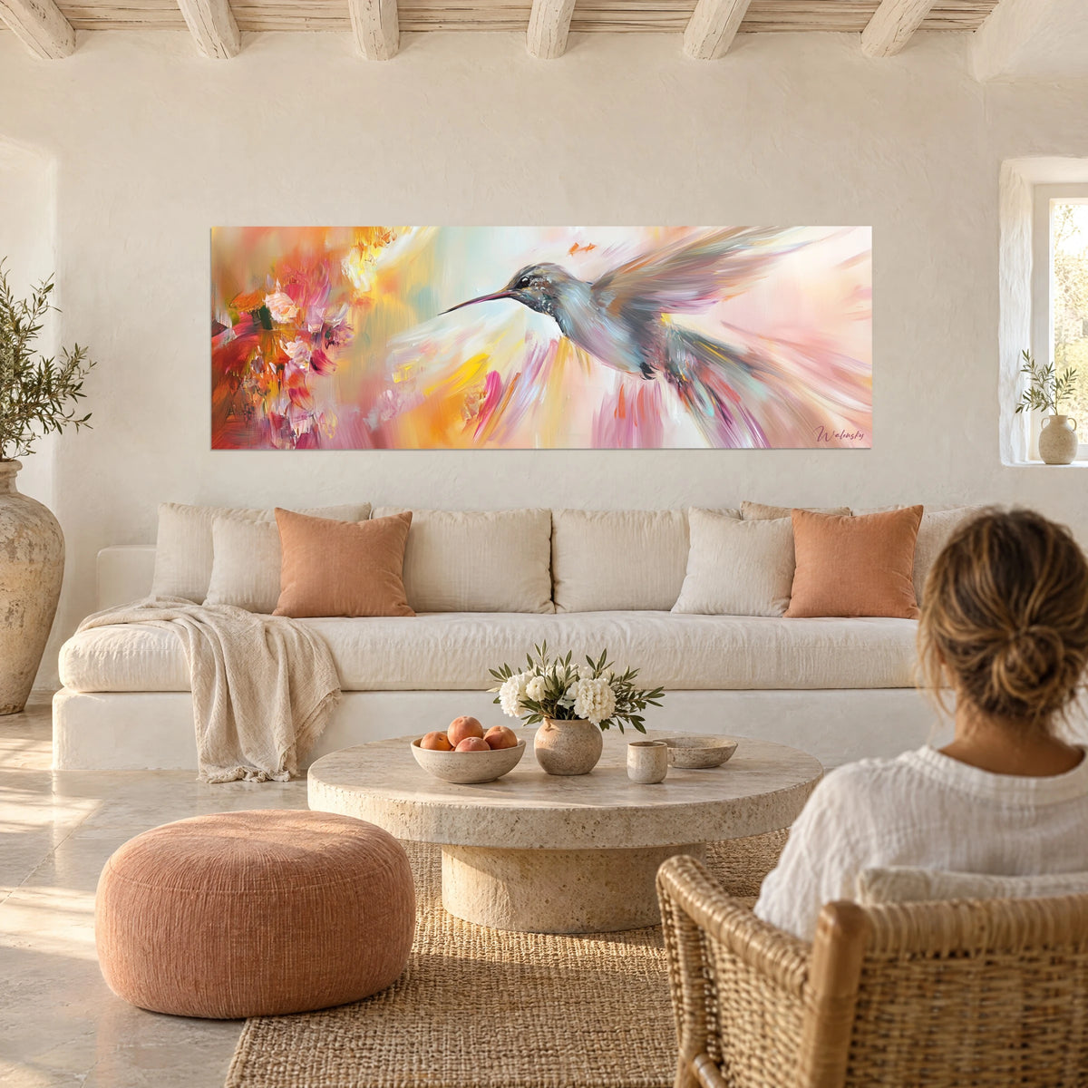 Wall art hummingbird with a long pink beak hovering within peach and yellow brushstrokes, above a sofa in a bright living room