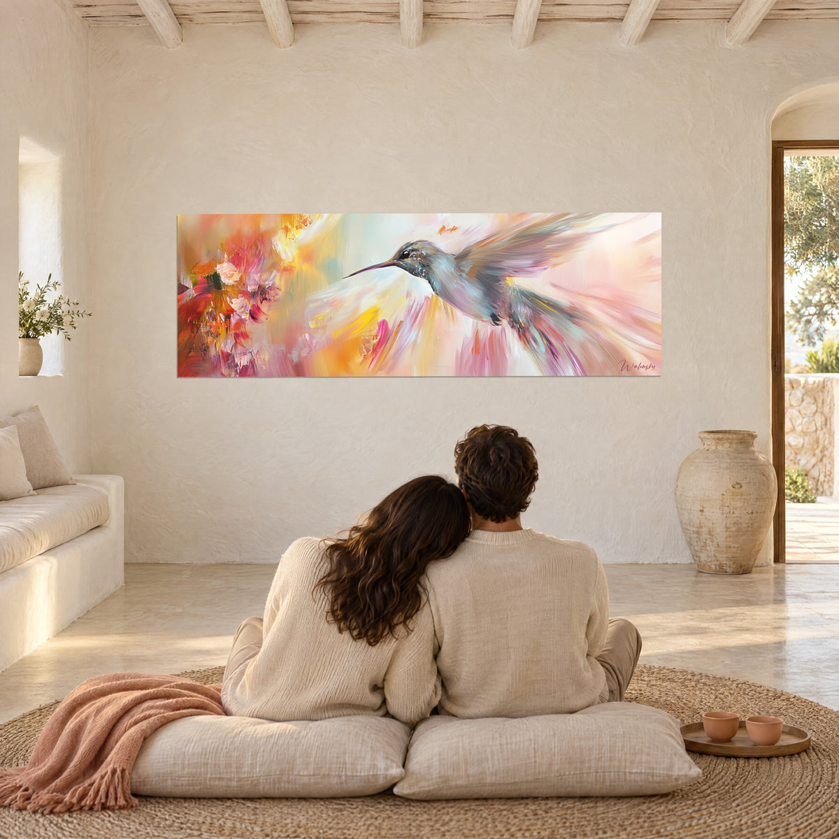 Wall art hummingbird sipping nectar in a cloud of pink, orange and yellow, admired by a couple seated back to the viewer on cream cushions