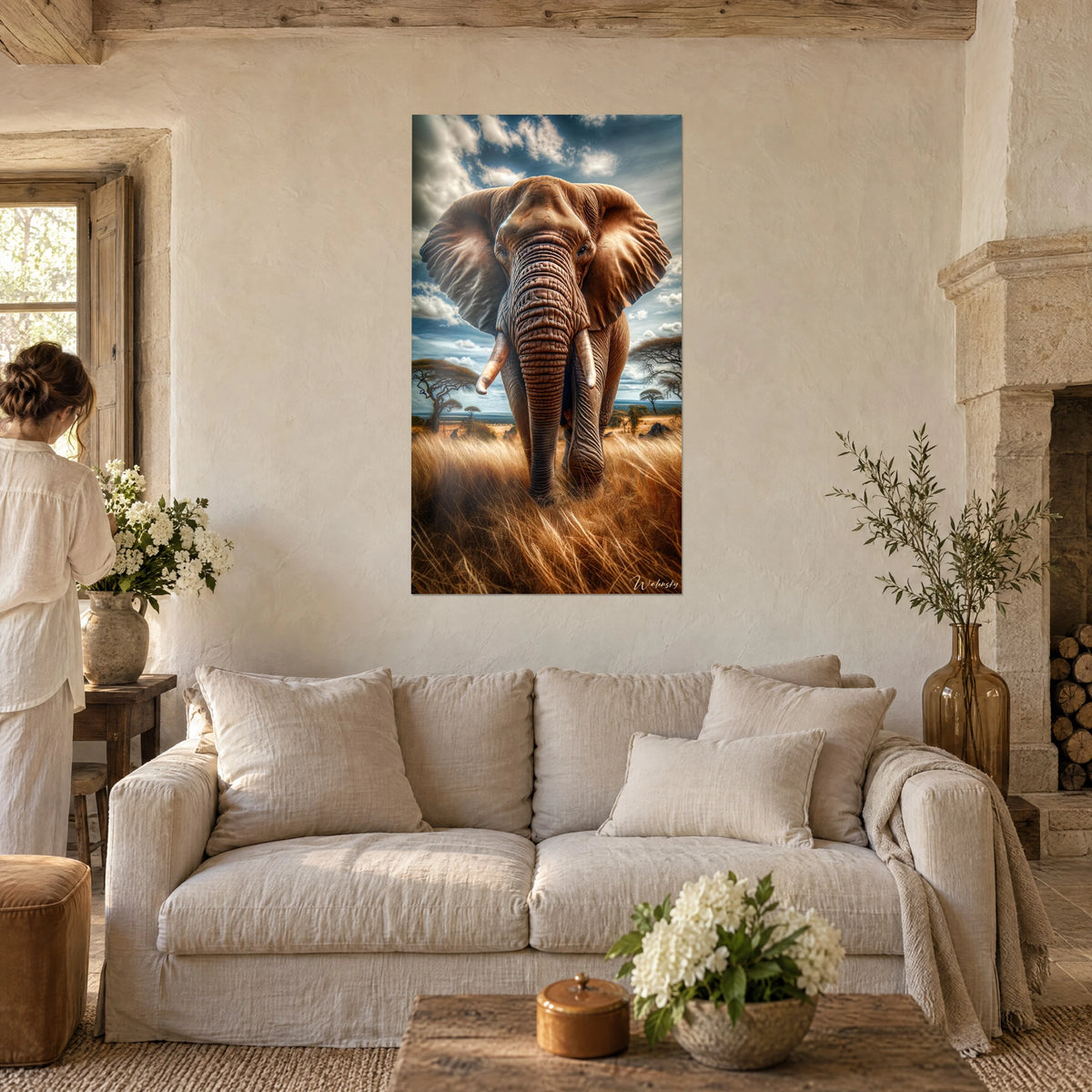 Schilderij van een Afrikaanse olifant met grote bruine oren, bewolkte blauwe lucht, vrouw van achteren, boven een bank in een lichte woonkamer