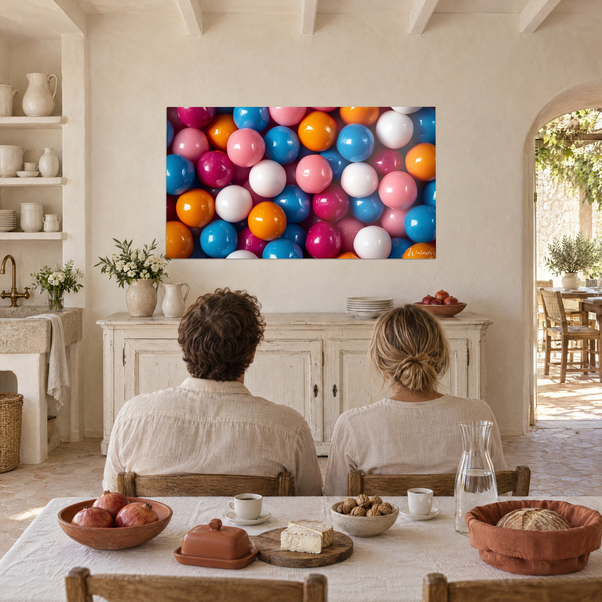 Cuadro de gomitas esféricas lacadas en blanco, magenta y azul celeste, dos personas de espaldas sentadas a una mesa en un mas