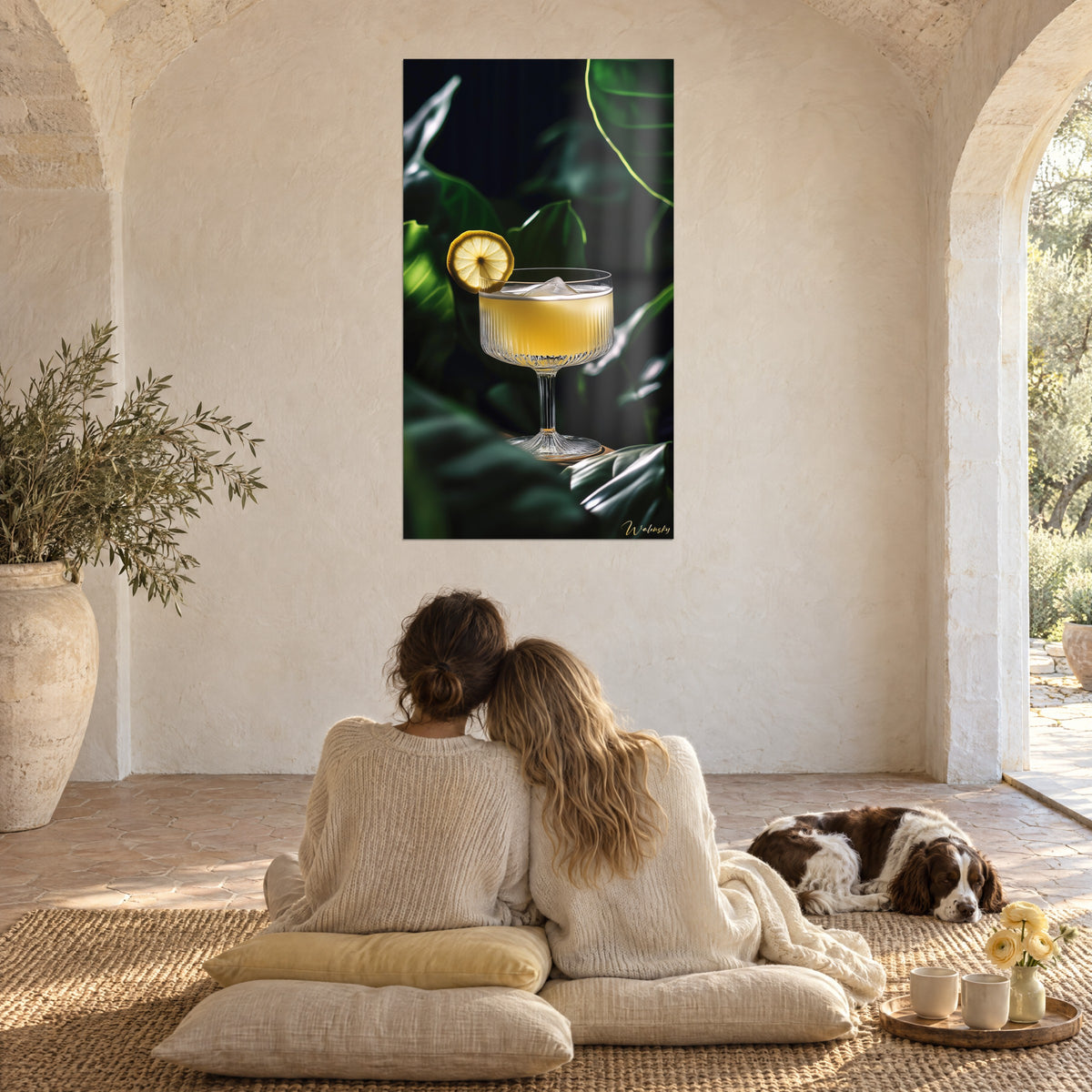 Tableau apéritif citronné servi dans une coupe cannelée, tranche dorée et feuillage noir-vert, mur chaux, couple de dos et spaniel