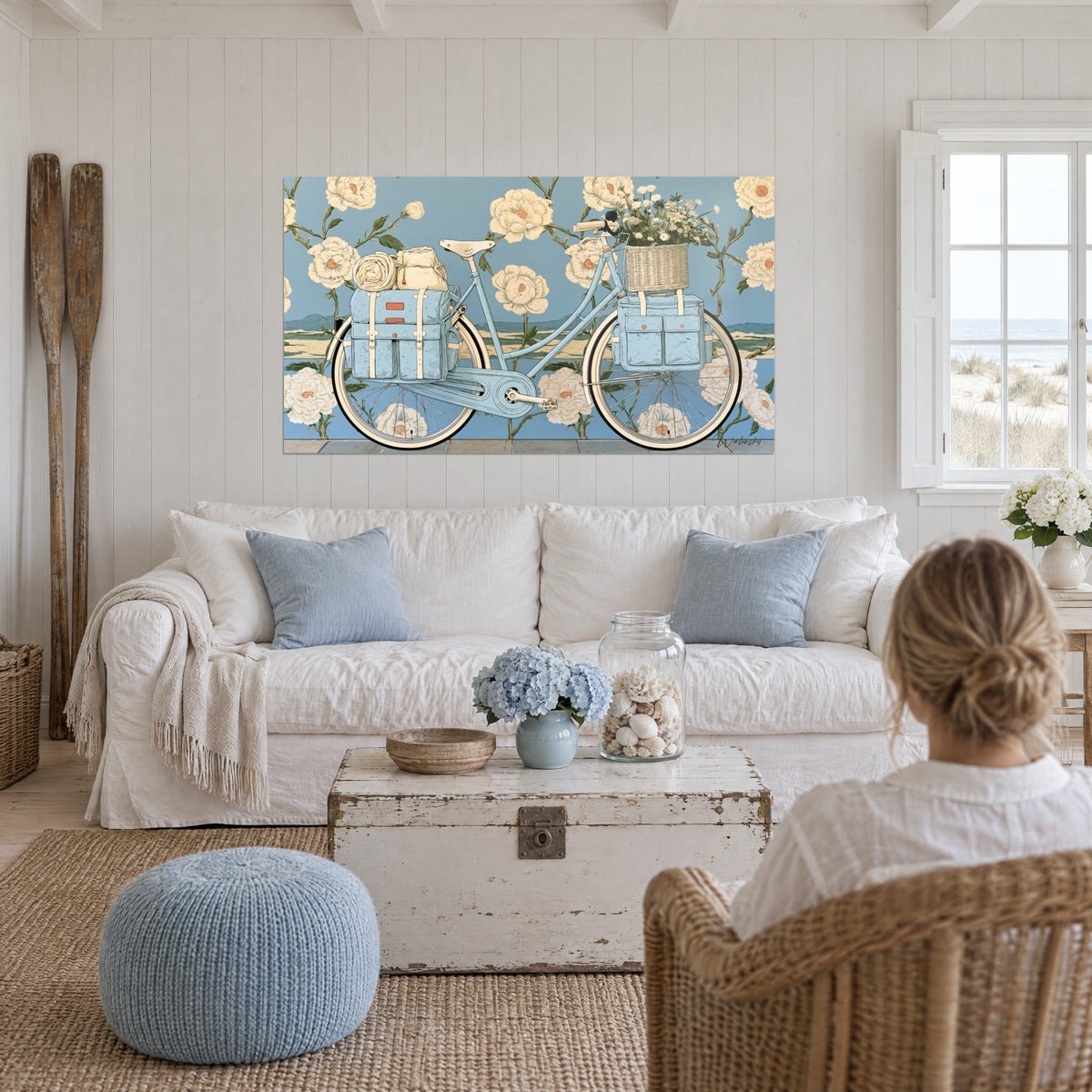 Tableau vélo bleu pastel chargé de sacoches crème devant des pivoines ivoire, au-dessus d'un canapé dans un salon lumineux