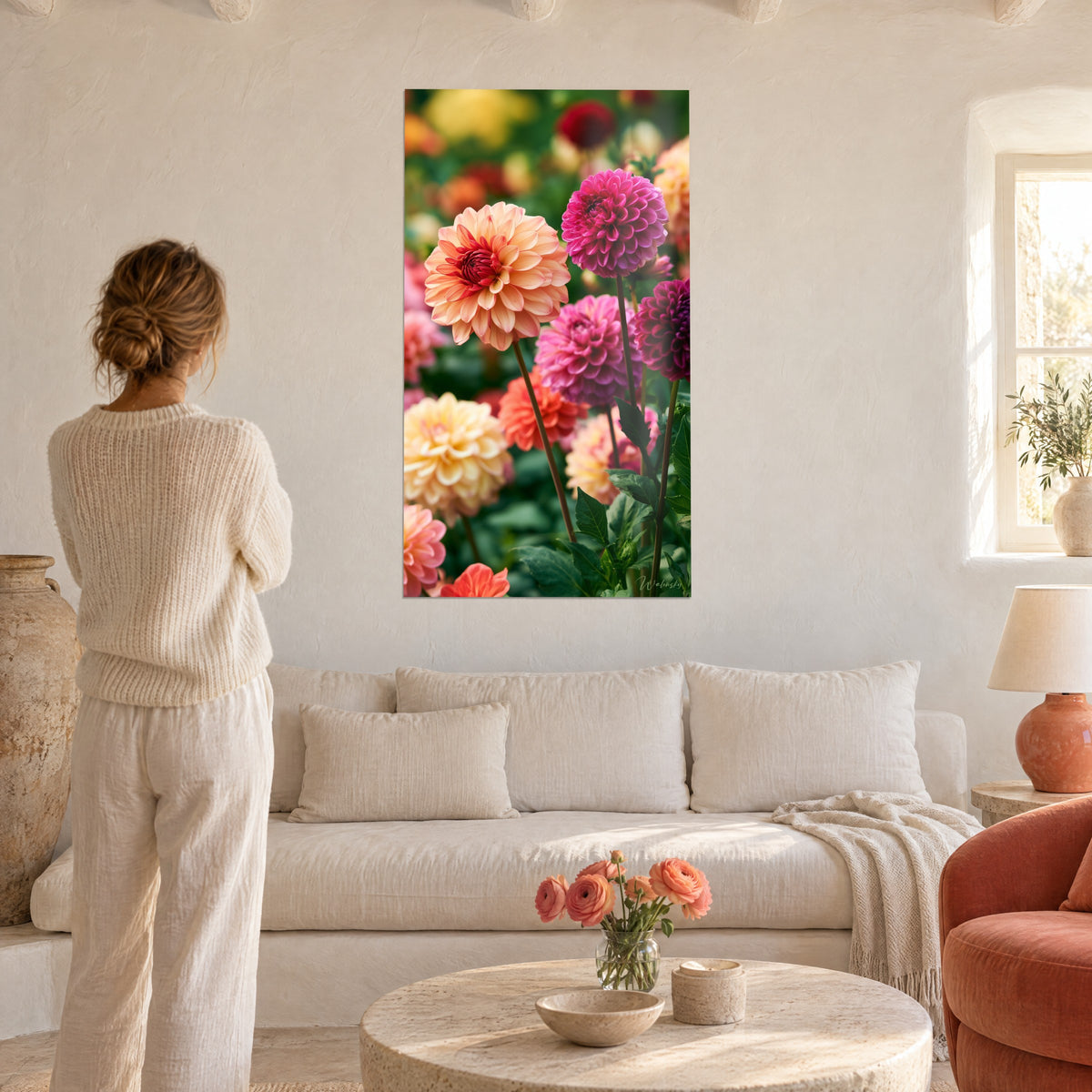 Wall art with peach and magenta dahlia blooms with a garnet-red heart, green foliage, woman seen from behind, above a sofa in a bright living room