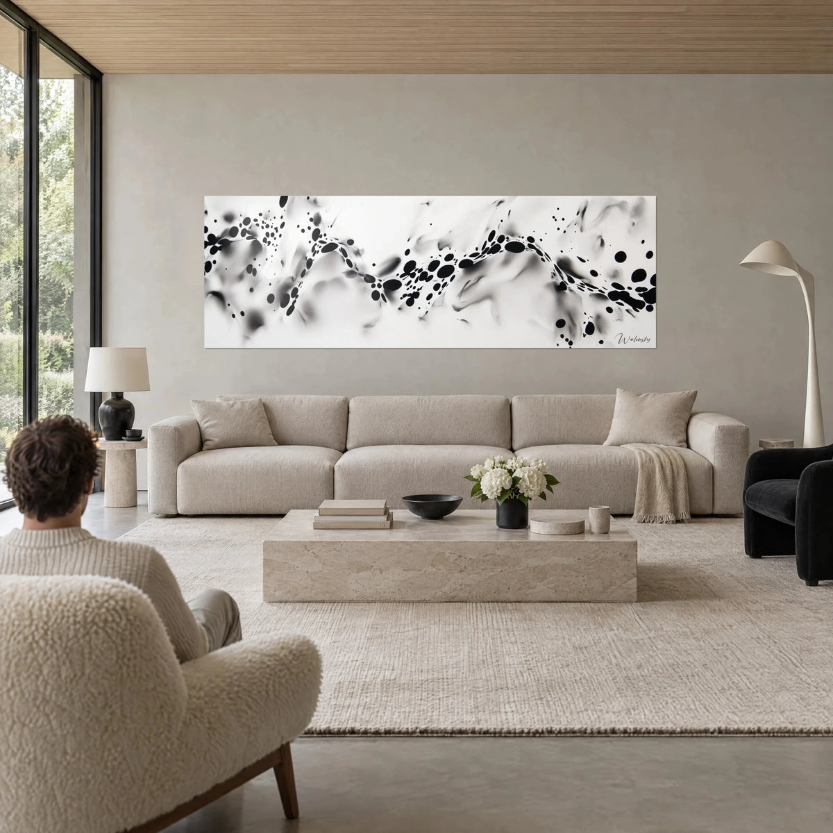 Schilderij lint van zwarte vlekken die golven op een witte ondergrond waarop grijze waas ligt, persoon van achteren boven een bank in een lichte woonkamer