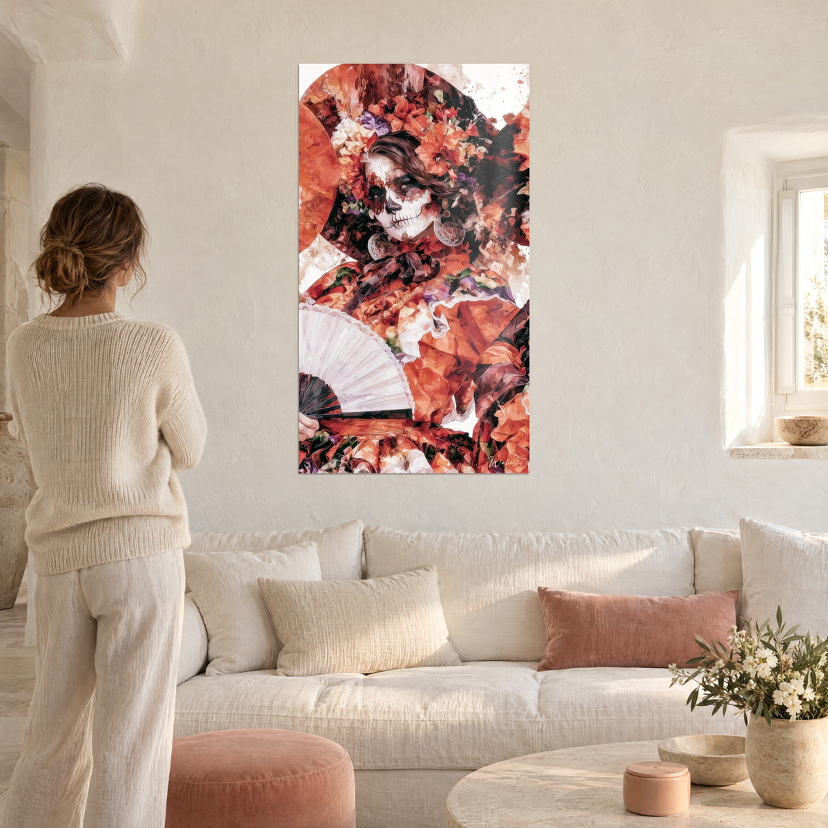Tableau visage de catrina peint blanc et noir, éventail ouvert et fleurs orange, femme de dos au-dessus d'un canapé dans un salon lumineux