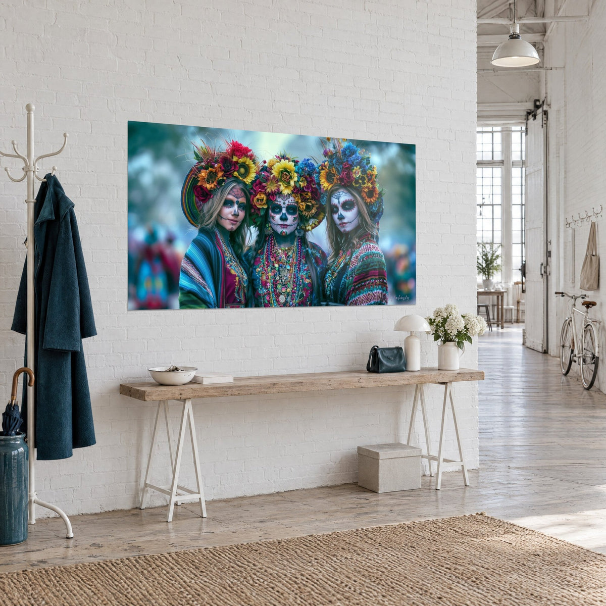 Tableau trio de catrinas fleuries, maquillage blanc et noir, ponchos rayés turquoise et rose, accroché de trois quarts sur un mur blanc de loft