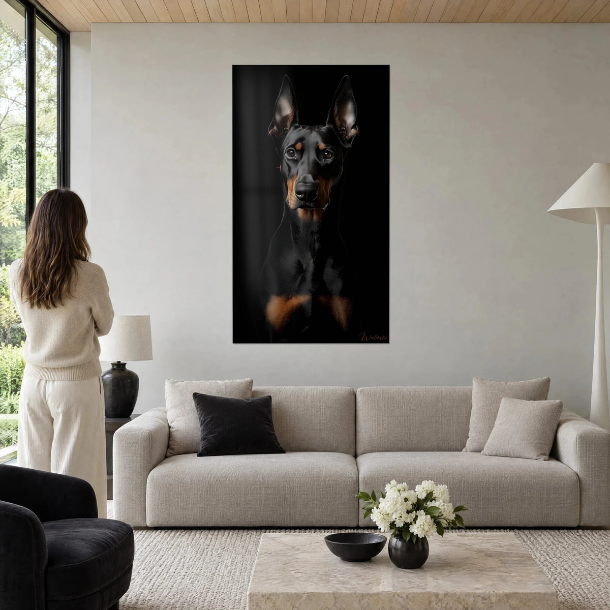 Tableau Doberman noir aux oreilles dressées, regard ambre sur fond sombre, femme de dos, au-dessus d'un canapé dans un salon lumineux