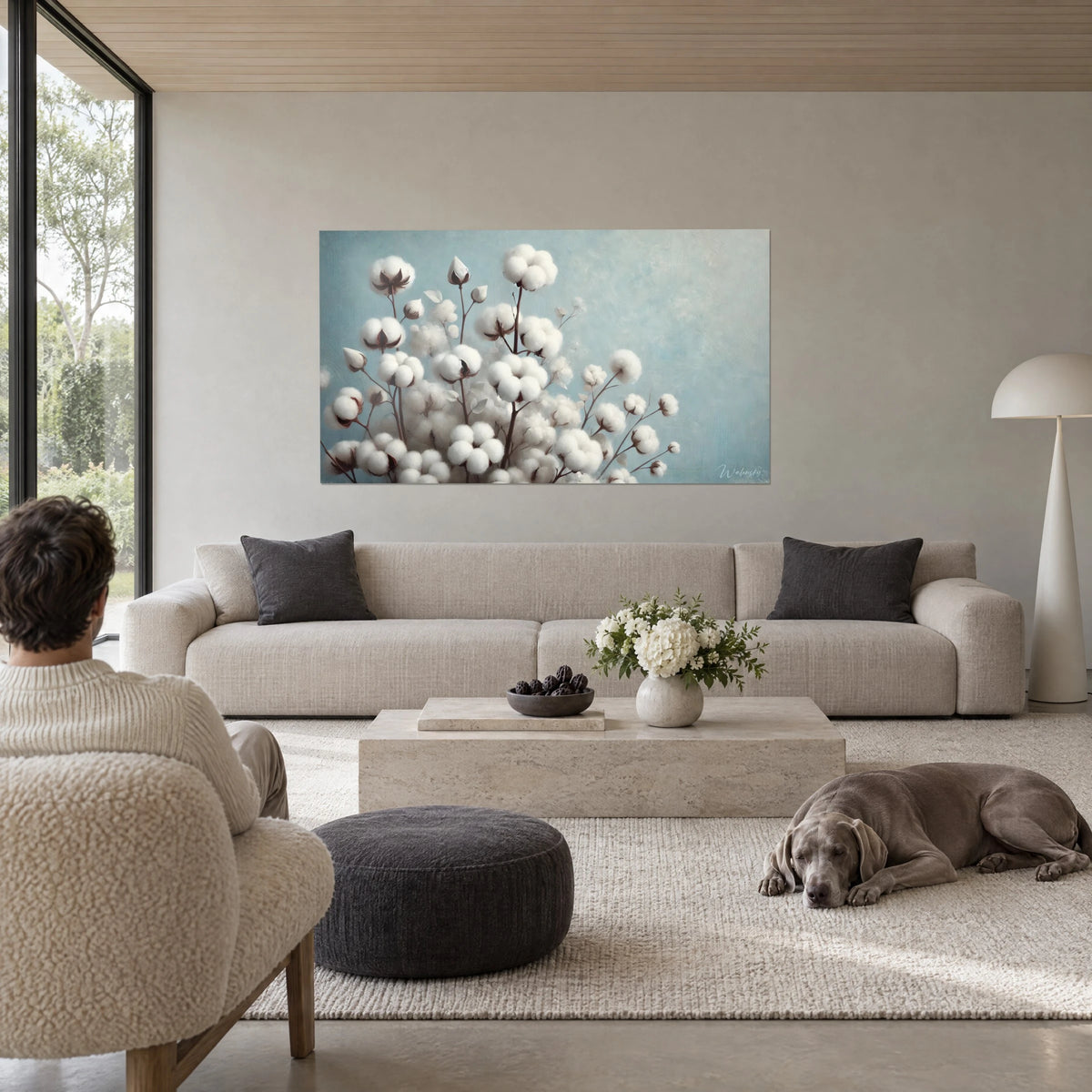 Cuadro ramo de flores de algodón blancas y mullidas, tallos marrón cacao sobre fondo azul cielo, persona de espaldas, encima de un sofá en un salón luminoso
