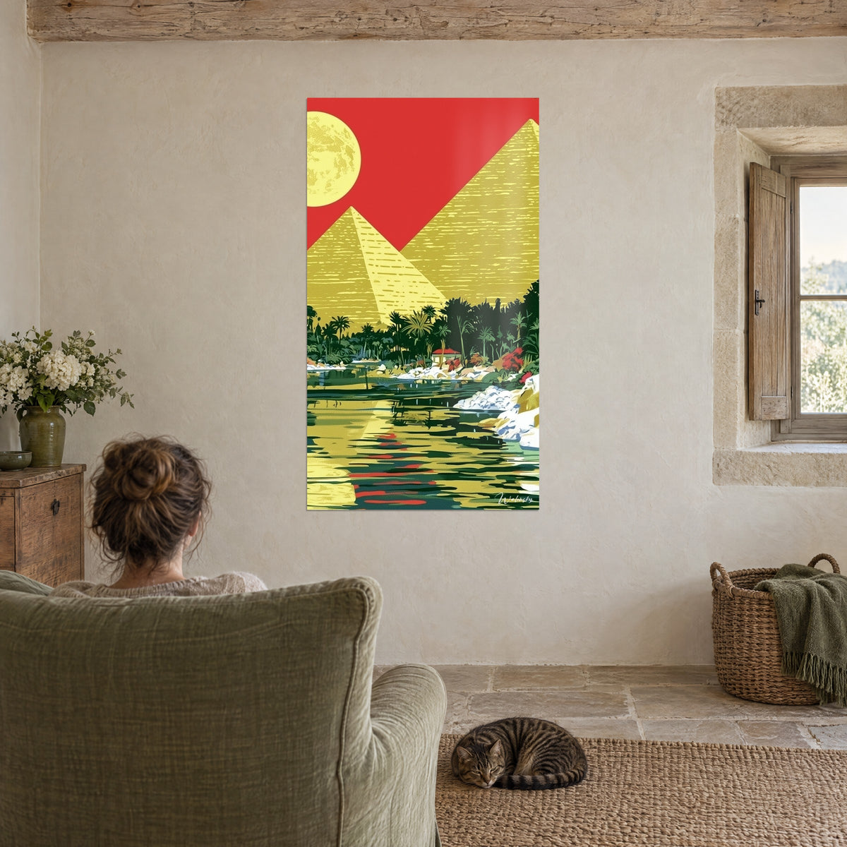 Quadro luna piena e piramidi in campiture gialle, cielo rosso e fiume verde, persona di spalle su una poltrona, gatto tigrato addormentato