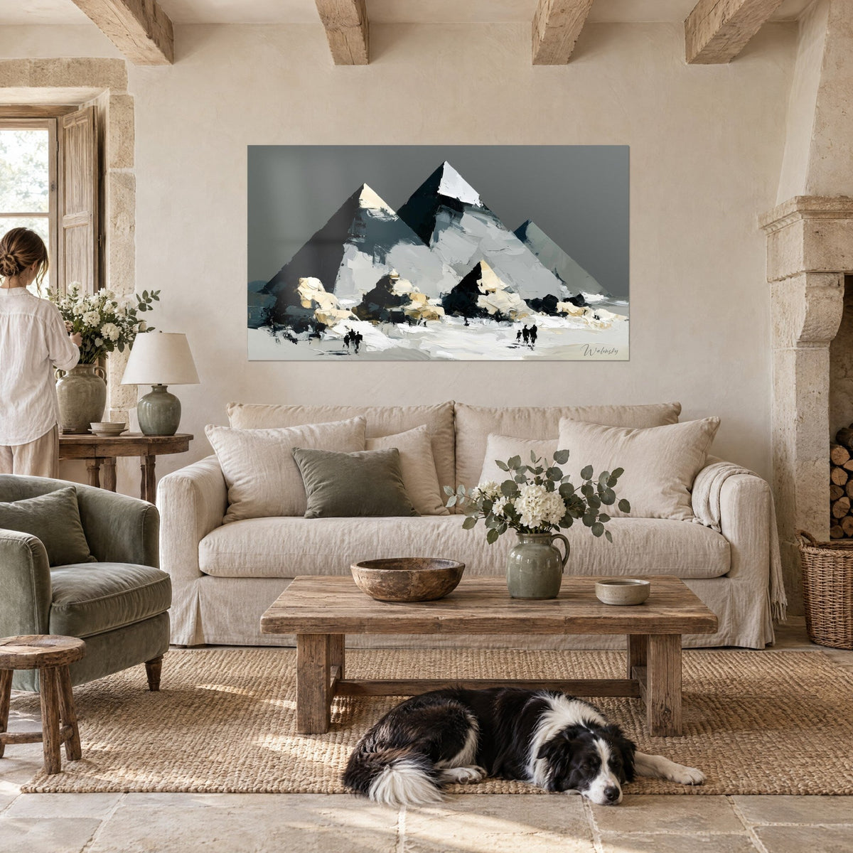 Wall art of grey and black pyramids in palette-knife painting, tiny silhouettes walking across white sand, above a sofa in a bright living room
