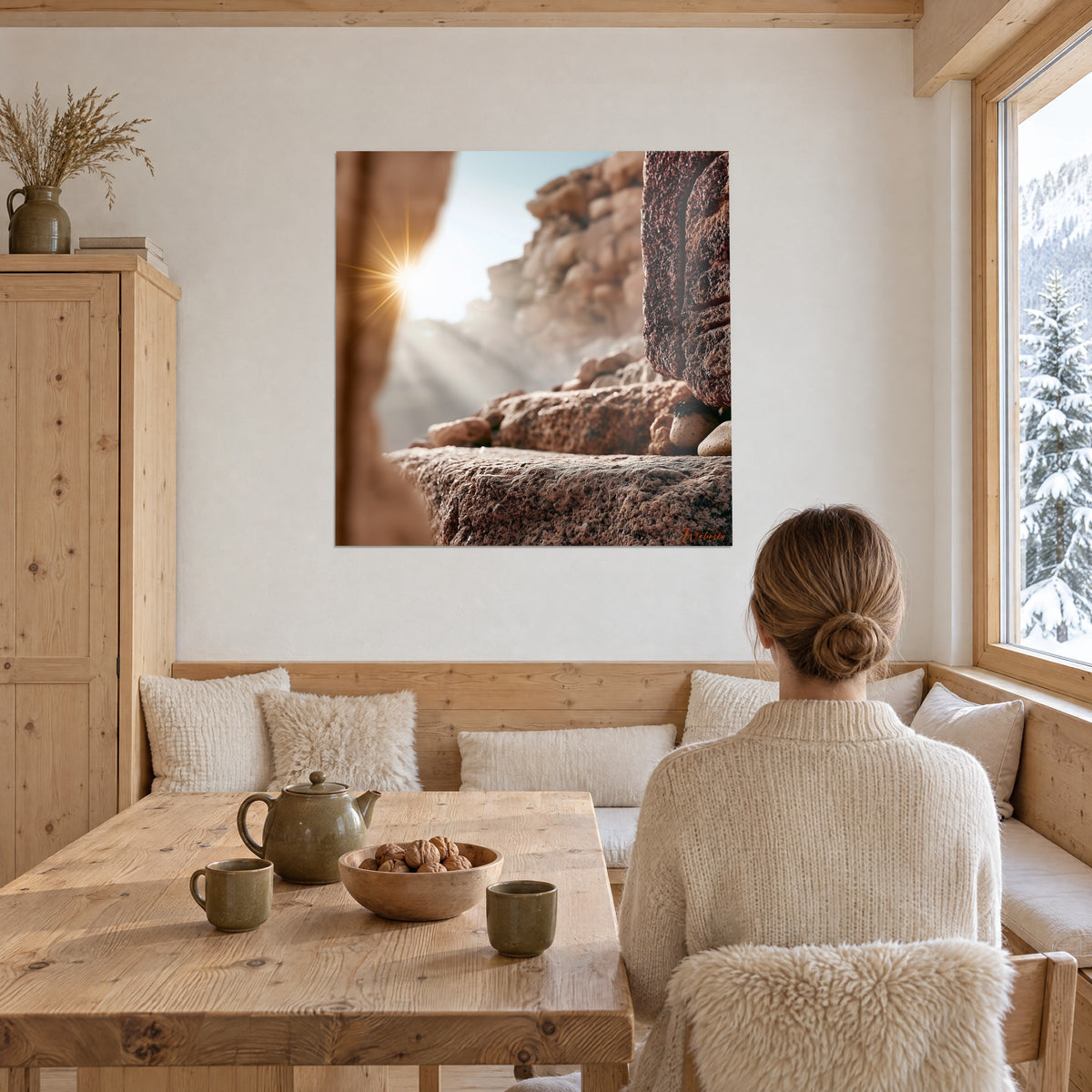 Gemälde einer Sandsteinschlucht mit Lichtstrahlen, beige Kiesel und rostfarbener Fels, Frau von hinten am Tisch neben einem Fenster mit Blick auf den Schnee des Chalets