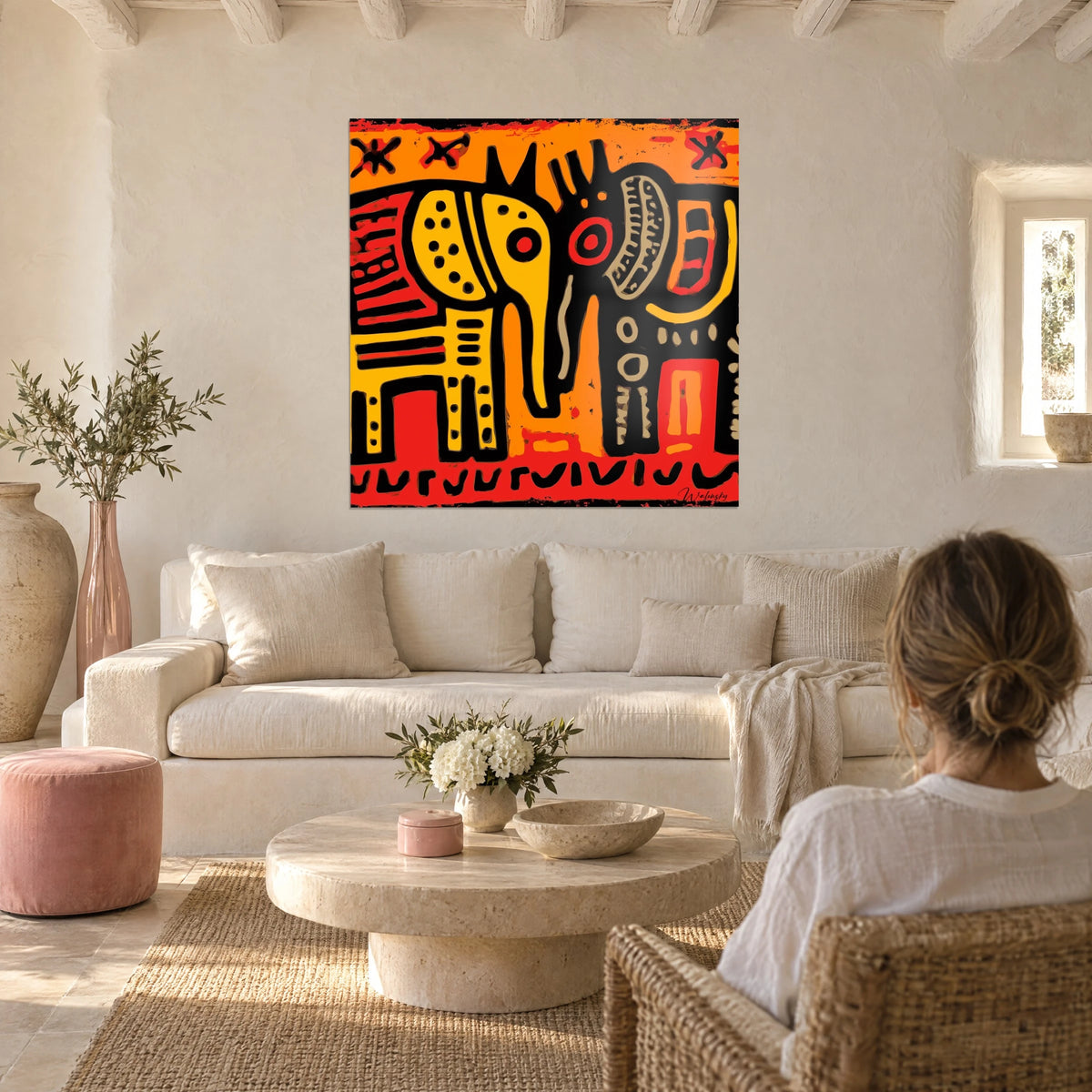Πίνακας με stylized ελέφαντα σε κίτρινο και κόκκινο, περιγραμμένο με μαύρο, φυλετικά μοτίβα, δύο άνθρωποι από πίσω πάνω από έναν καναπέ σε ένα φωτεινό σαλόνι