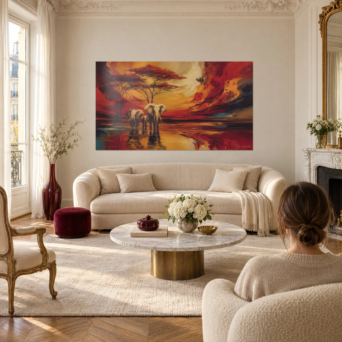 Schilderij drie Afrikaanse olifanten in een rood en goudkleurig meer, acacia's met karmijnrode kruinen, een vrouw van achteren, boven een bank in een lichte woonkamer