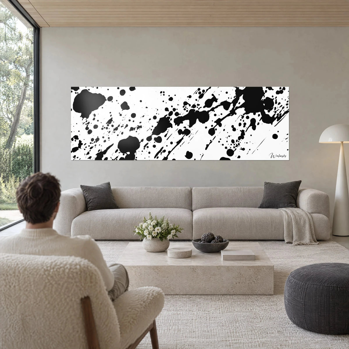Gemälde mit schwarzen Tintenspritzern auf weißem Grund, diagonal verlaufende Tropfen, ein Mann von hinten über einem Sofa in einem hellen Wohnzimmer