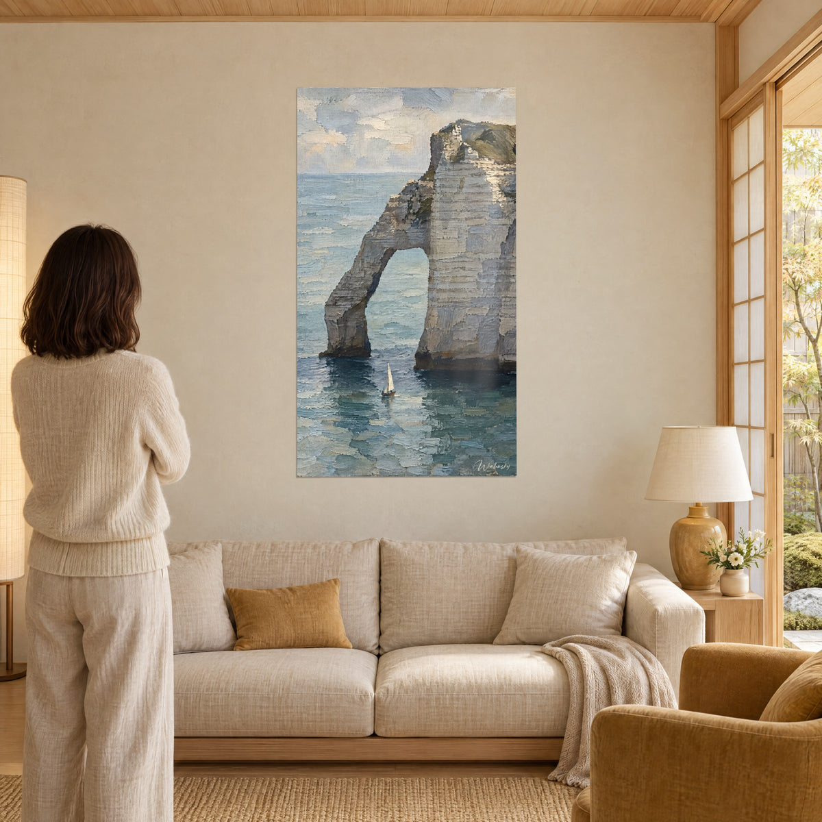 Schilderij krijtboog van Étretat boven blauwgroene zee, minuscule witte zeilboot, een vrouw van achteren, boven een bank in een lichte woonkamer